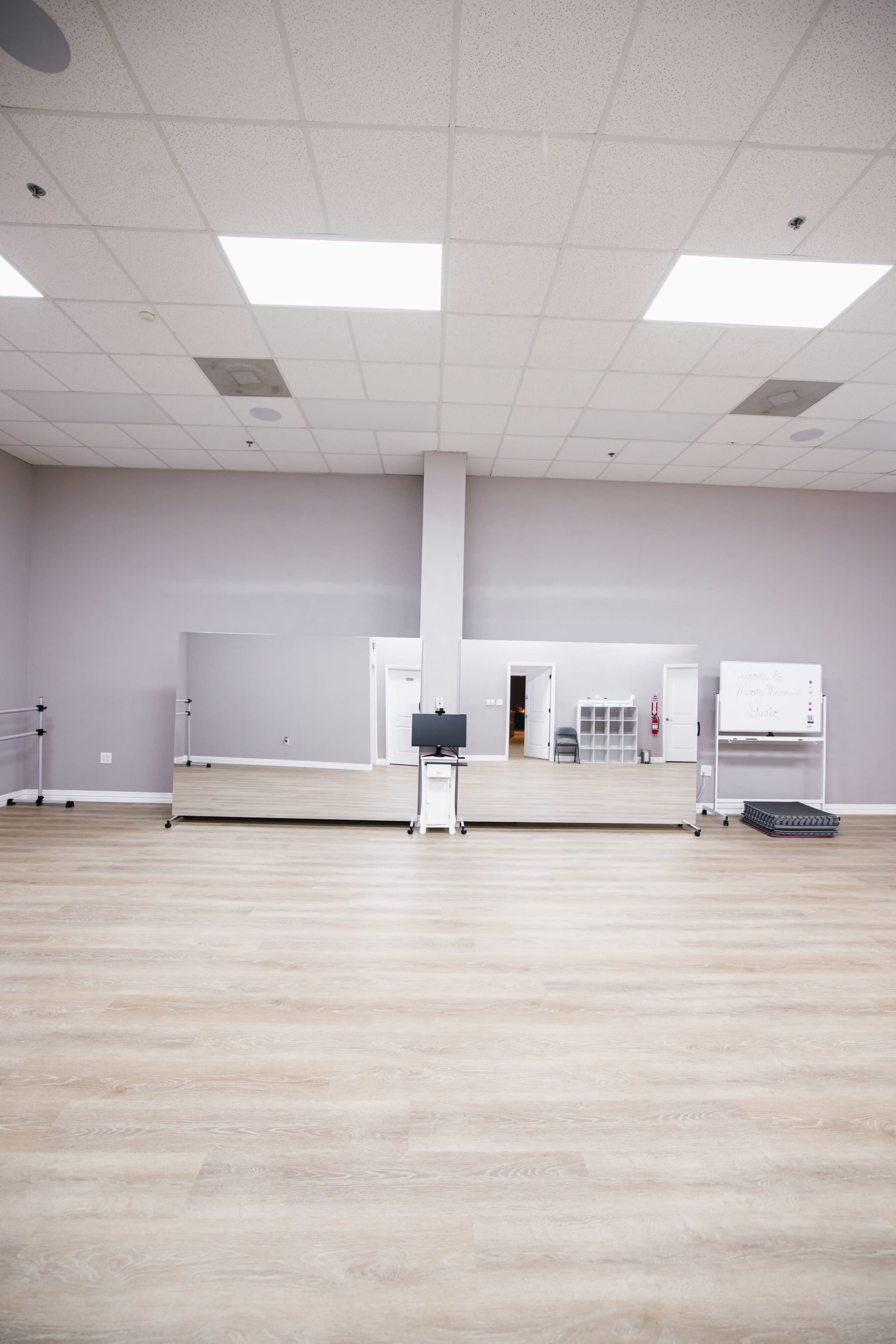 A spacious dance studio features a large wall mirror, light-colored wooden flooring, and minimal furnishings against a neutral wall.
