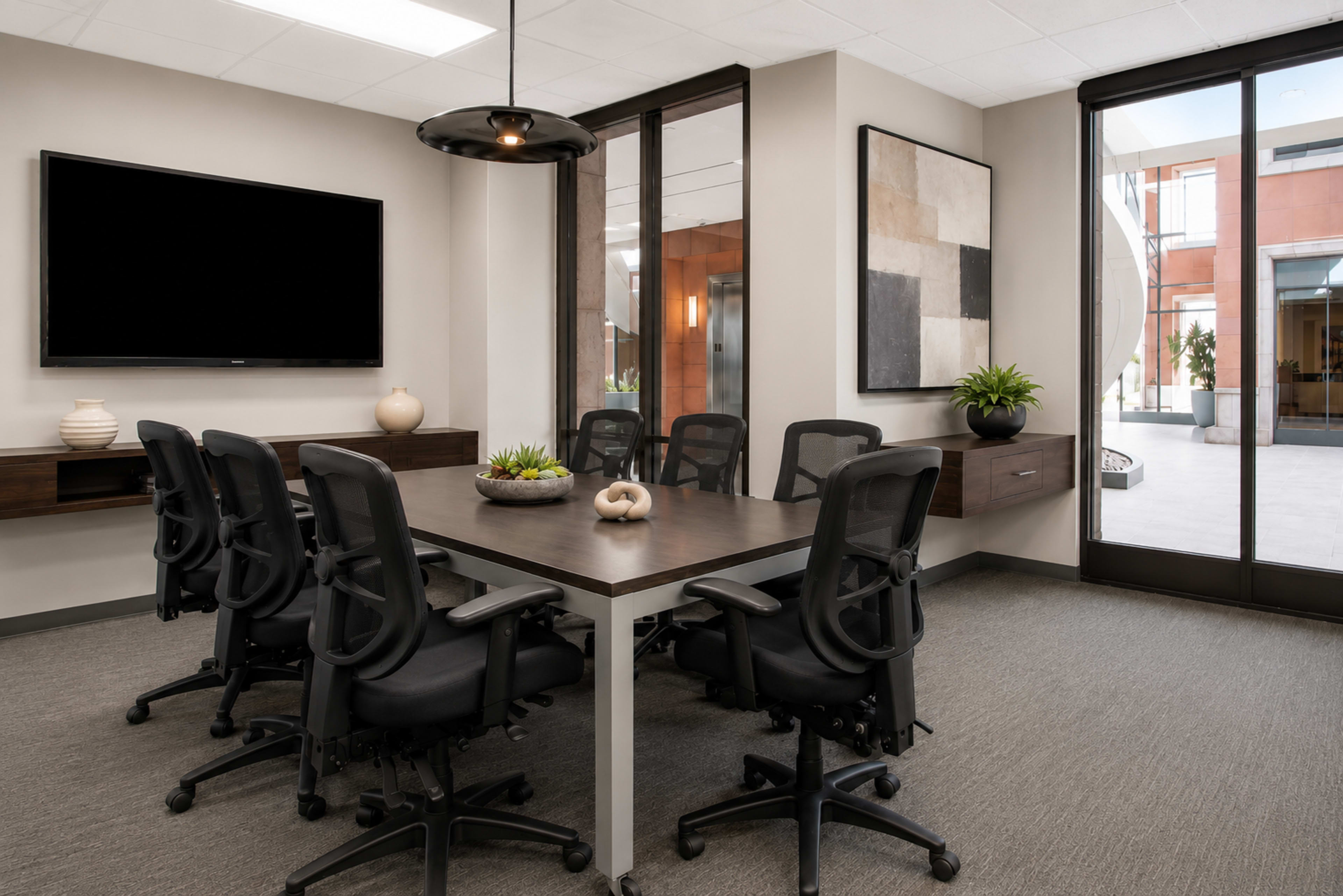 A modern conference room features a large table surrounded by ergonomic chairs, with a TV mounted on the wall and large windows allowing natural light to enter.