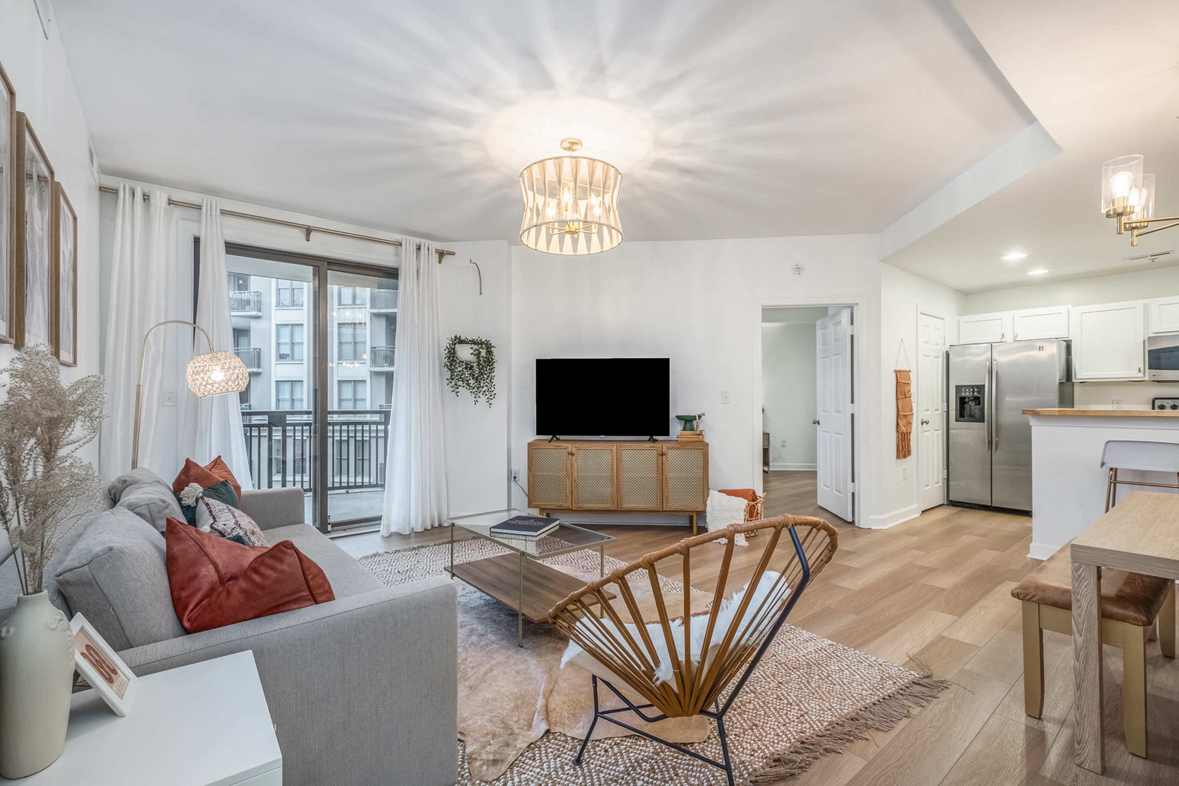 The living room features a modern design with a television on a wooden cabinet, a gray sofa with decorative pillows, and a balcony visible through sliding glass doors.