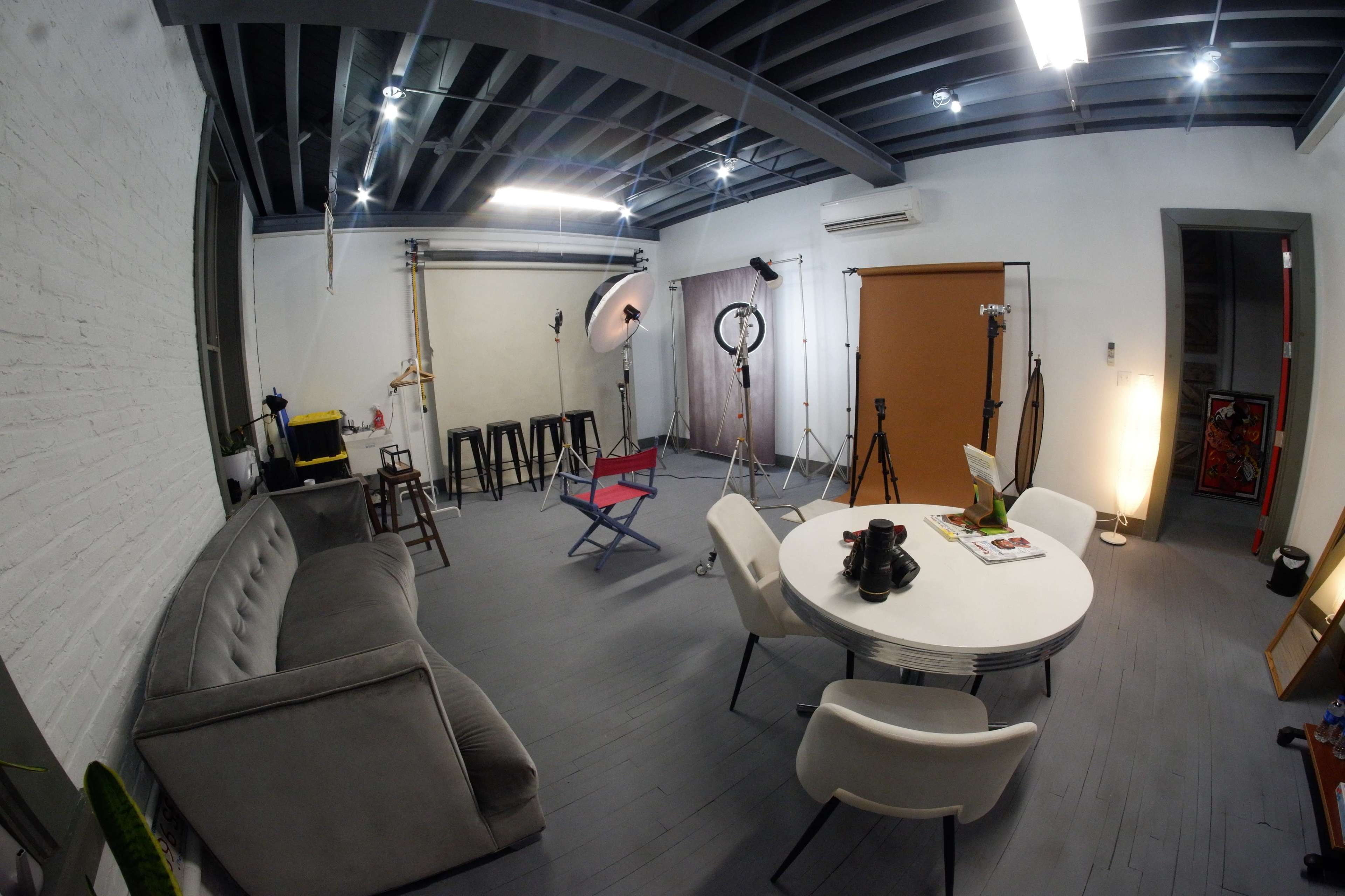 The image shows a studio space equipped with lighting equipment, a round table, chairs, and a gray couch, with softbox lights and backdrops arranged for a photo shoot.
