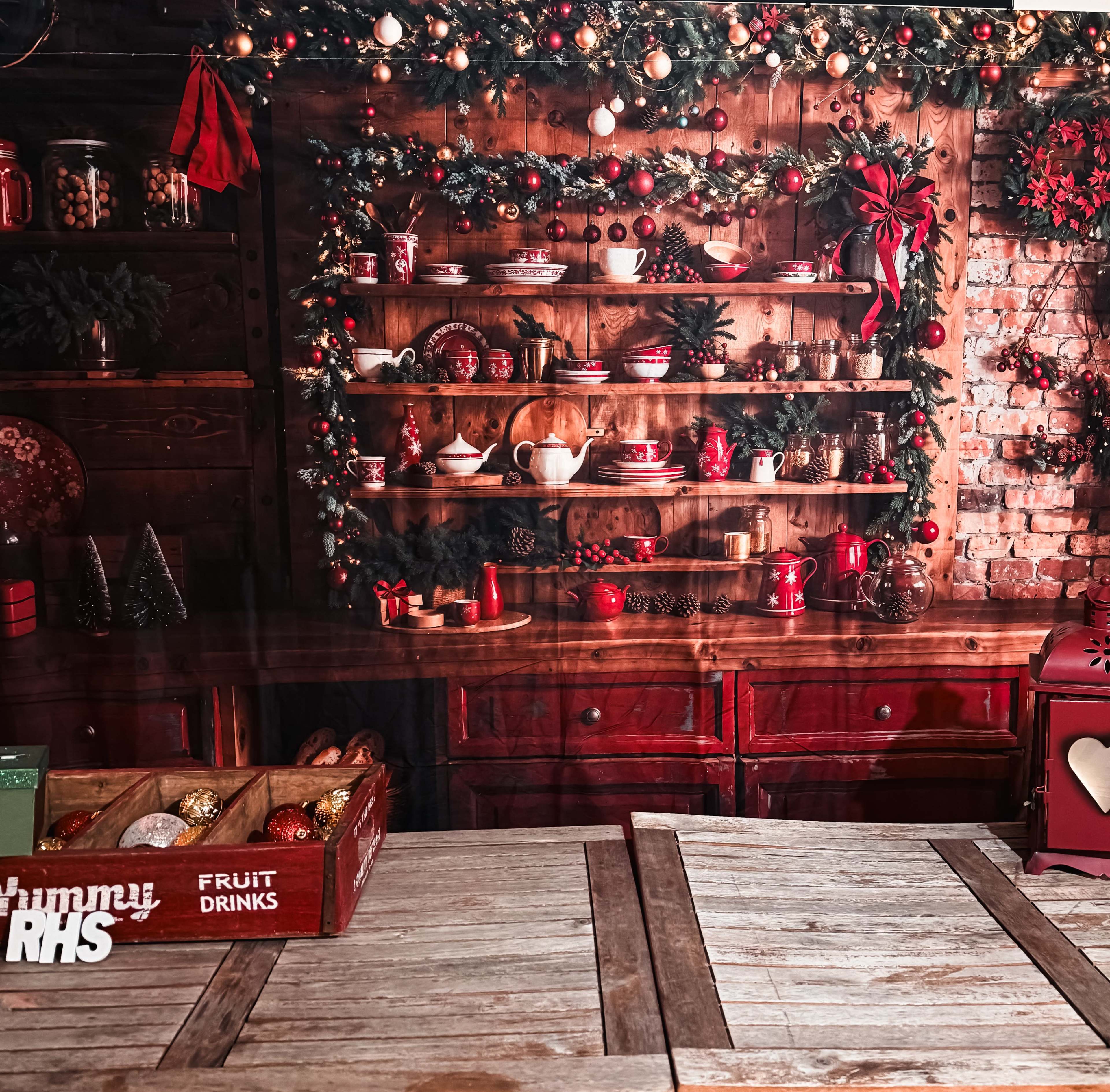 The image shows a festive wooden shelf adorned with holiday decorations, including red and white dishware, surrounded by garlands and ornaments, against a rustic brick wall.