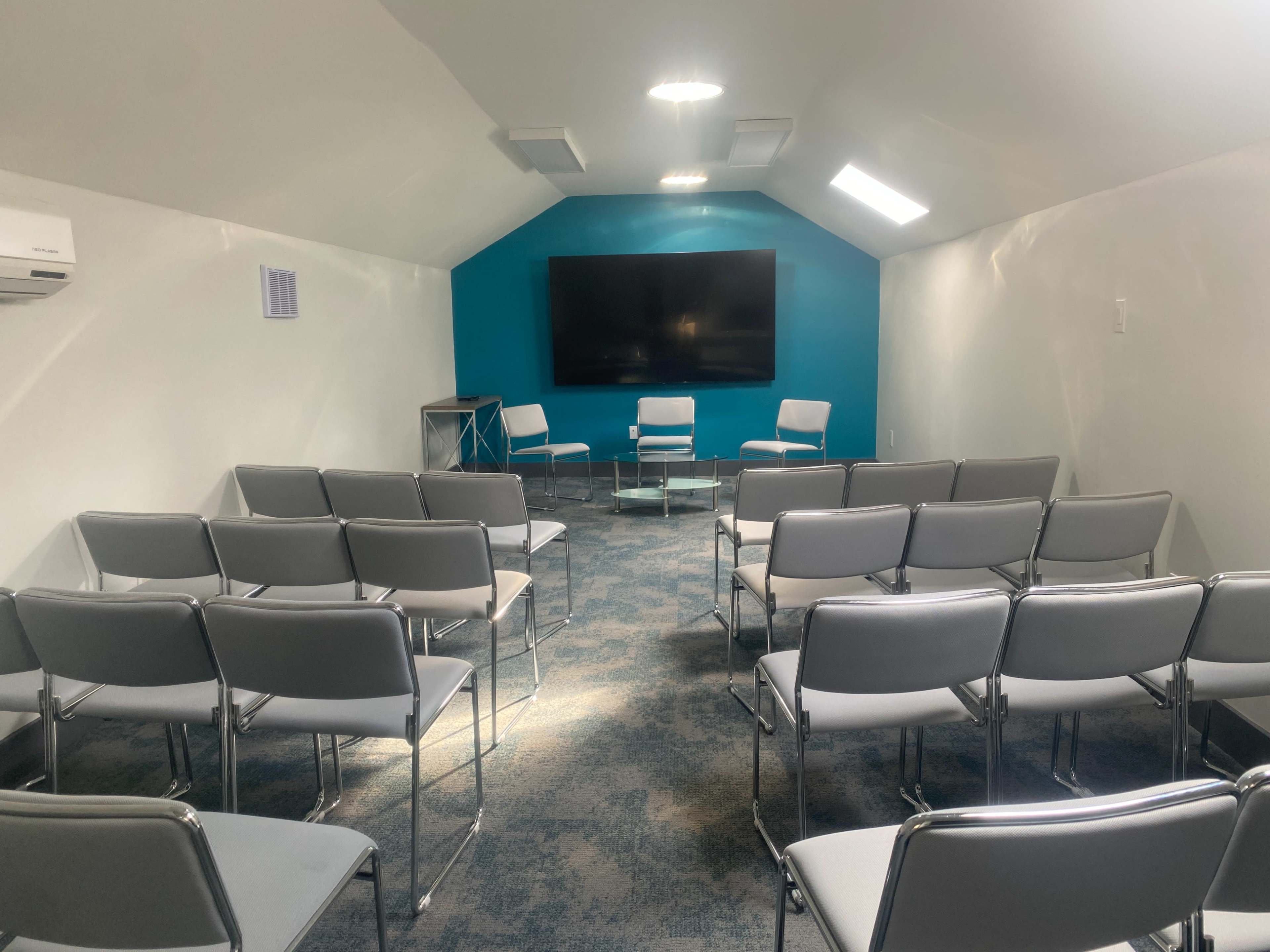 A small meeting room features an arrangement of gray chairs facing a large screen on a blue accent wall.
