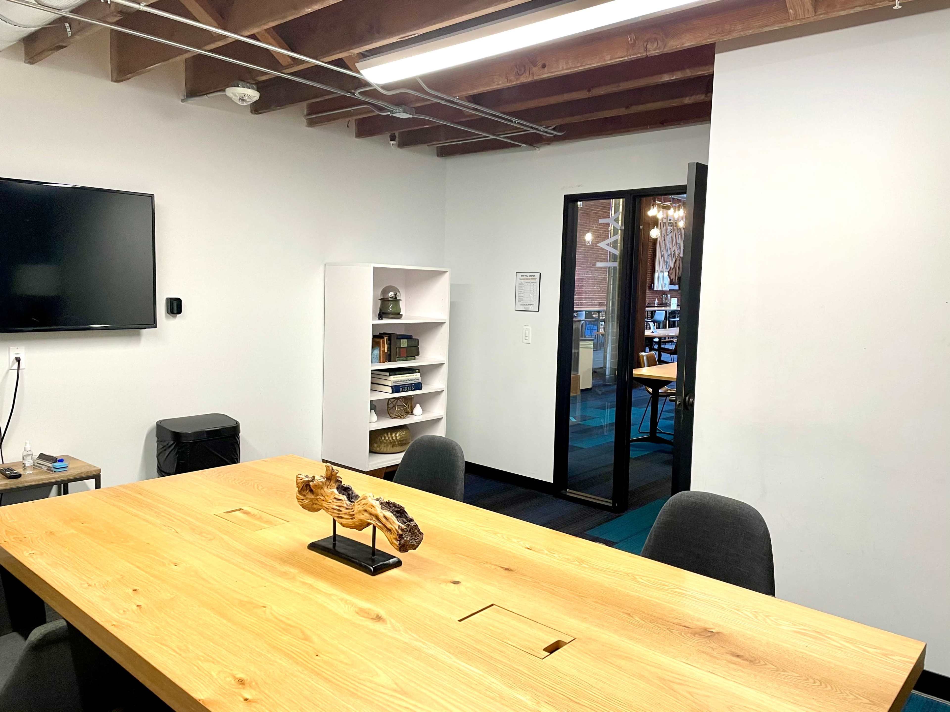 A wooden conference table with a centerpiece sits in a well-lit room, featuring a TV on the wall and a glass door leading to another workspace.