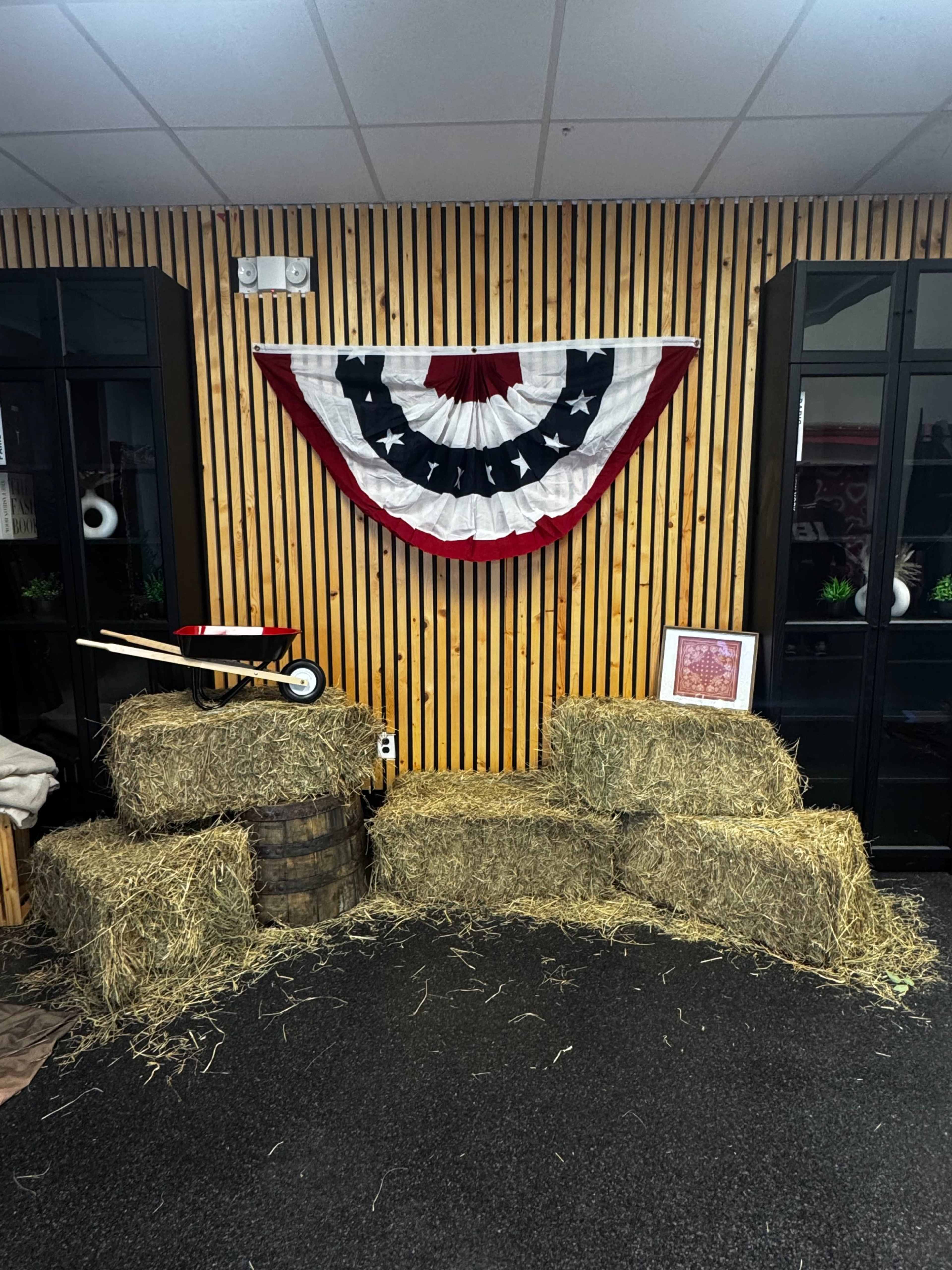 The scene features a wall adorned with a red, white, and blue bunting, surrounded by bales of hay and a wooden barrel, set against a backdrop of vertical wooden slats.
