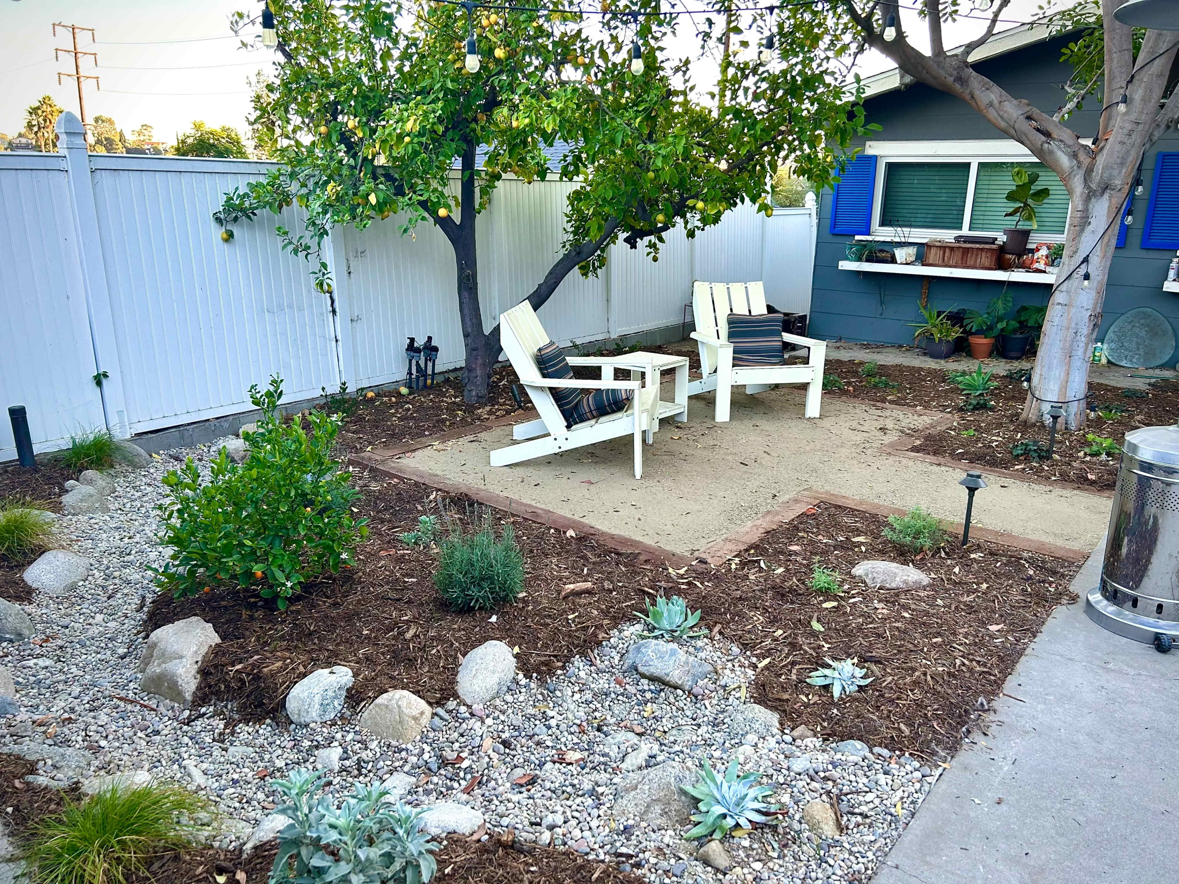 A landscaped backyard features two white chairs on a sandy area surrounded by gravel, rocks, and various plants, with a tree and a house in the background.