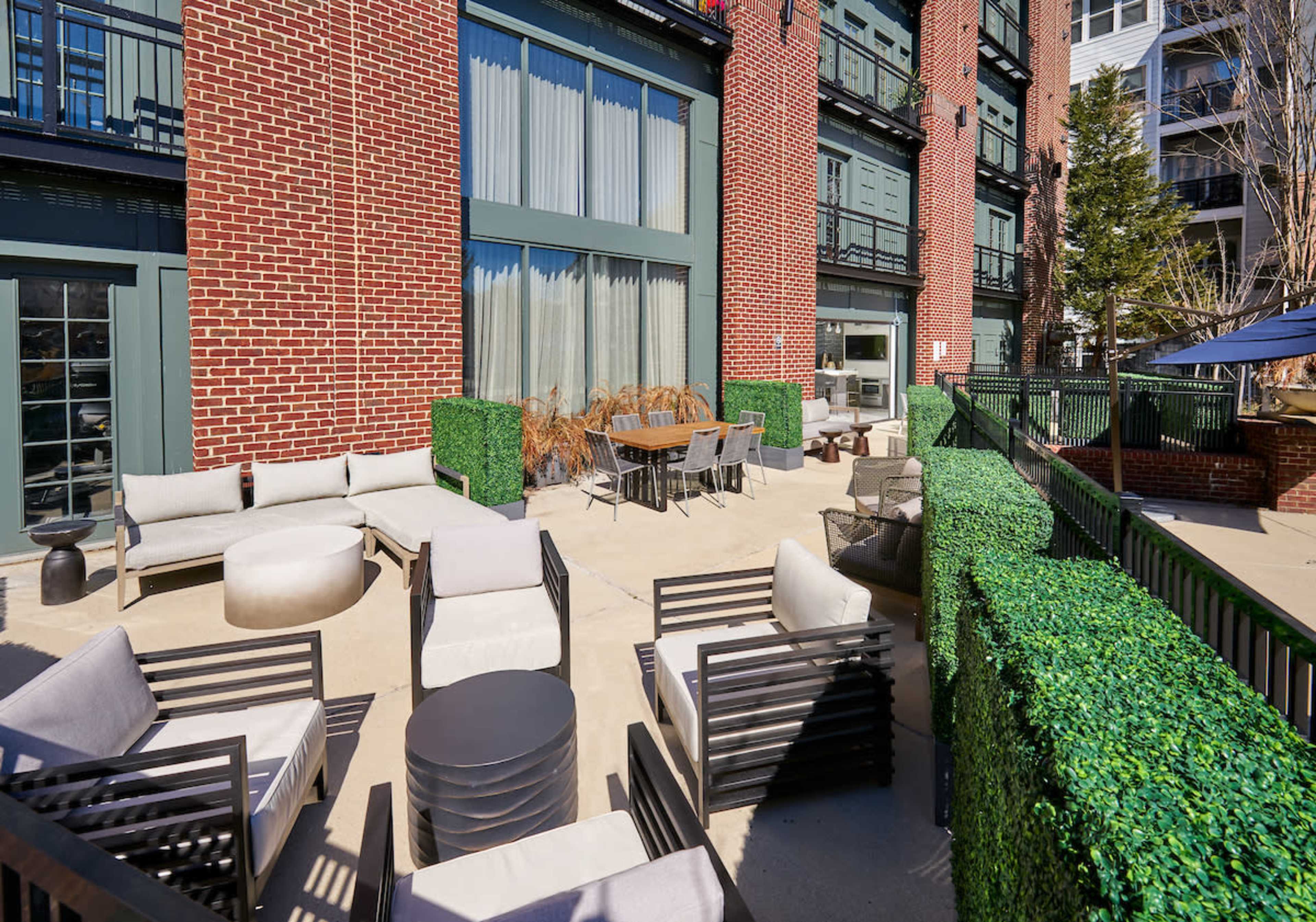 A landscaped outdoor patio features furniture arrangements, including sofas, a circular table, and potted plants, adjacent to a brick building.