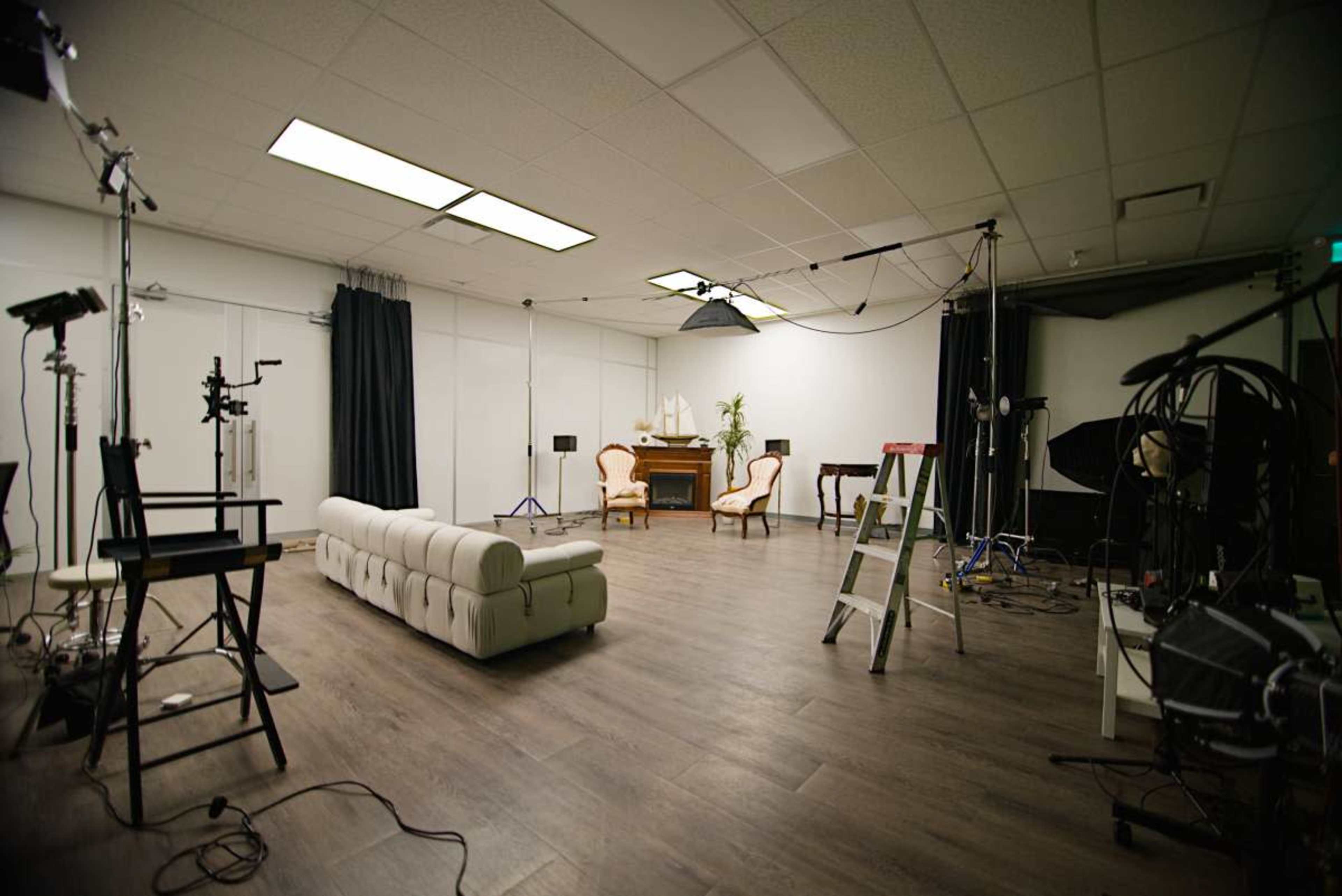The image shows a spacious studio with a light-colored floor, a white couch, two chairs, and various lighting equipment set up for a photoshoot.