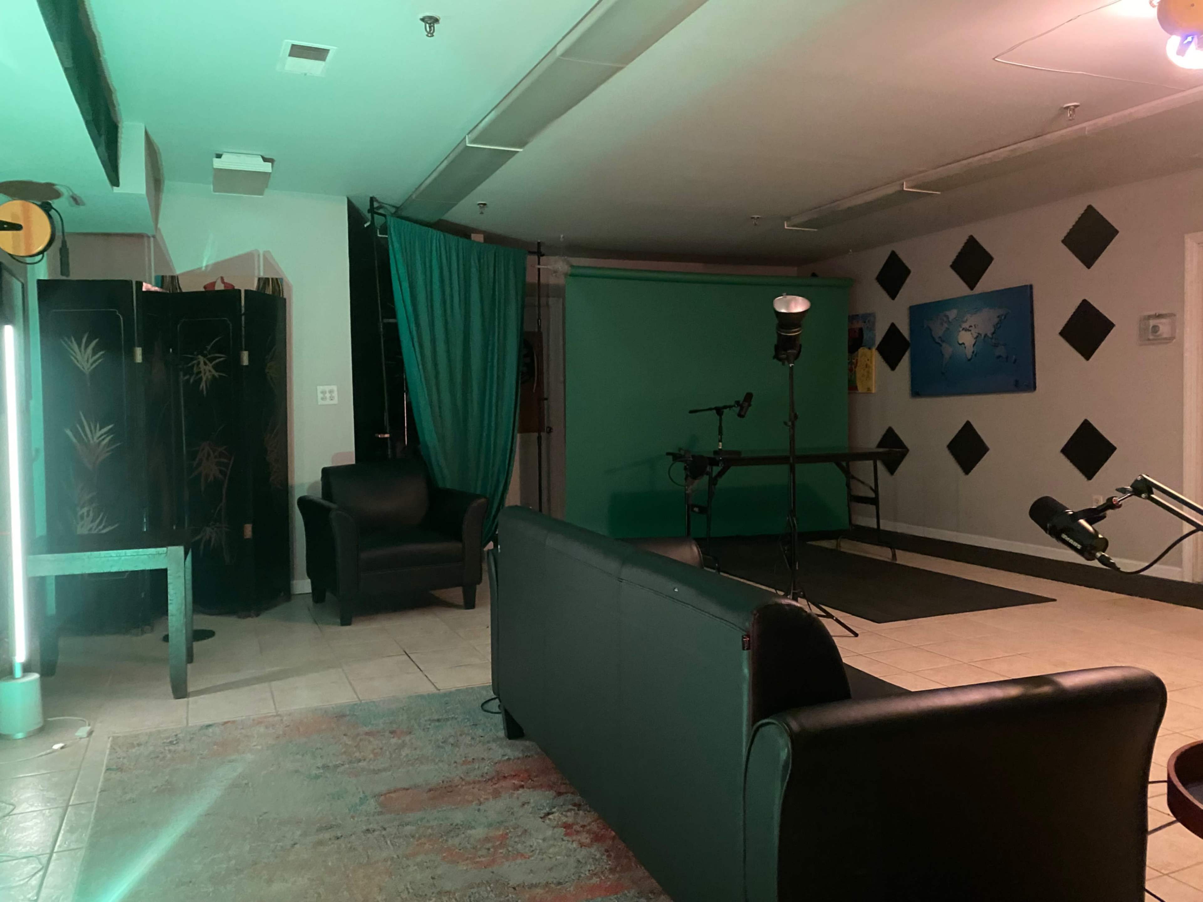 A dimly lit room with a green backdrop, a black sofa, and a microphone setup, indicating a space for video or audio production.
