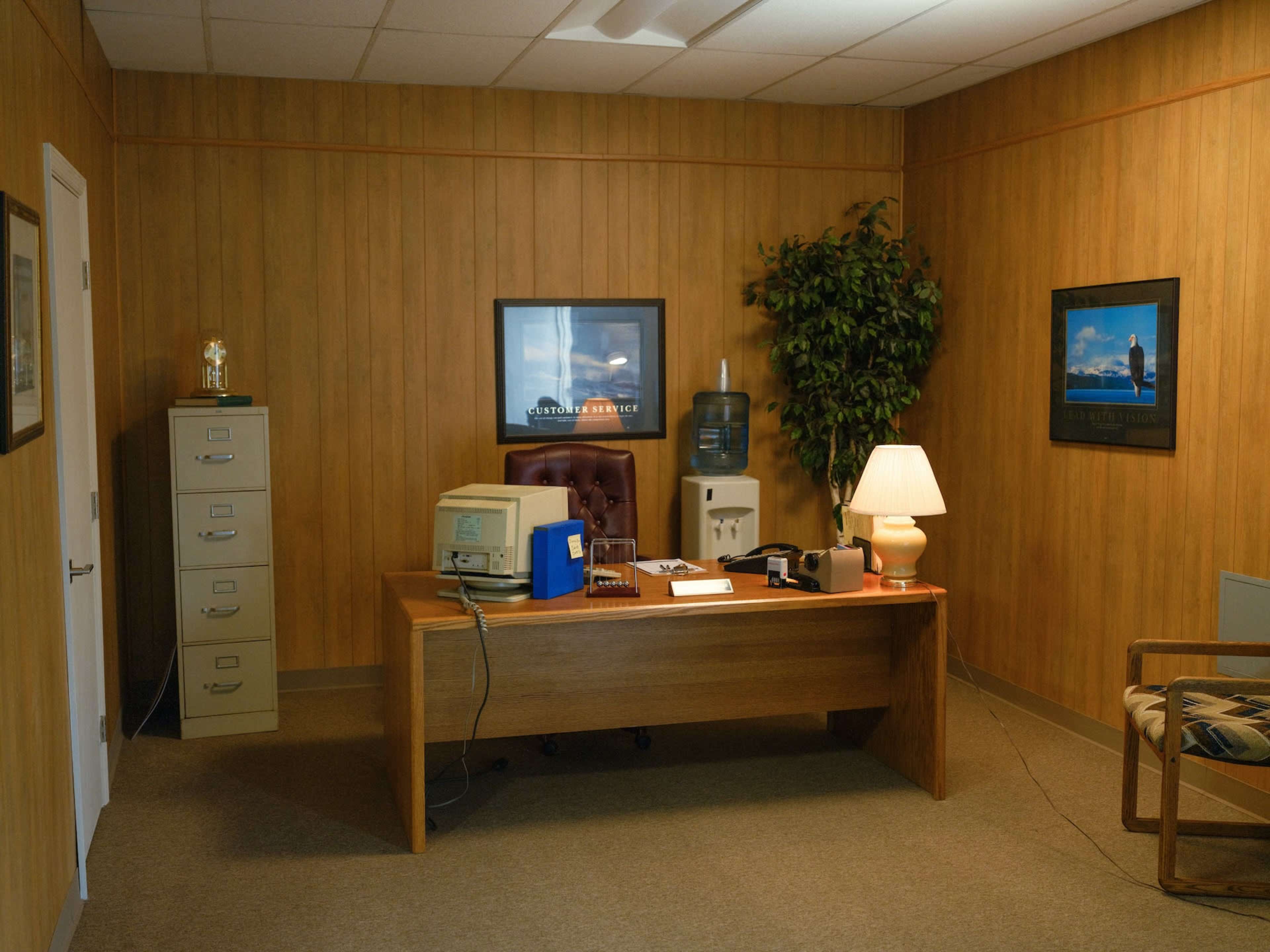 The image shows a wooden-paneled office with a desk, a vintage computer, a filing cabinet, and a water cooler, along with framed pictures on the wall.