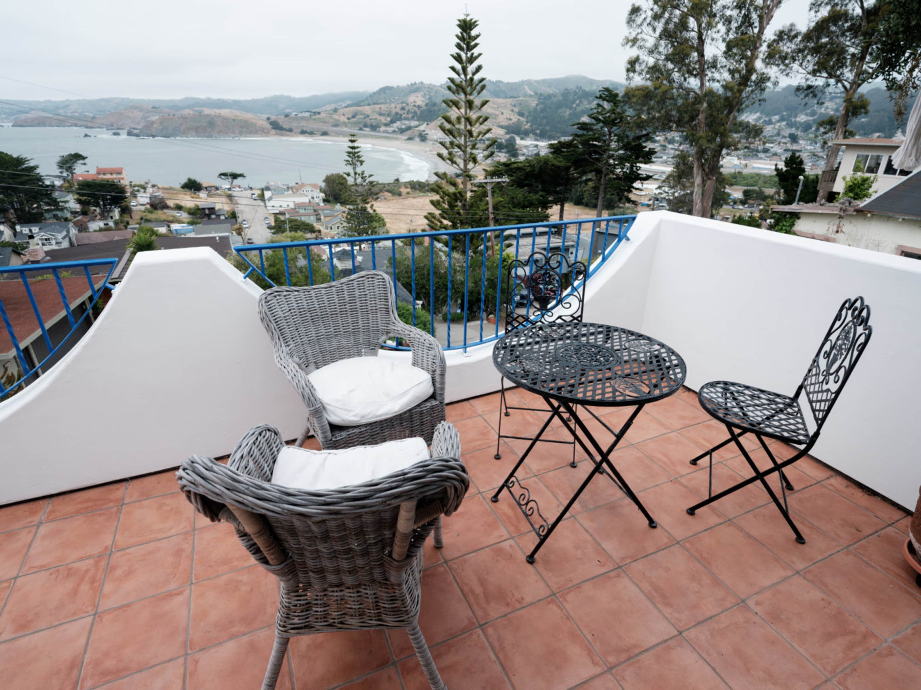 A small balcony features a round metal table and two chairs, with a view of a coastal landscape in the background.