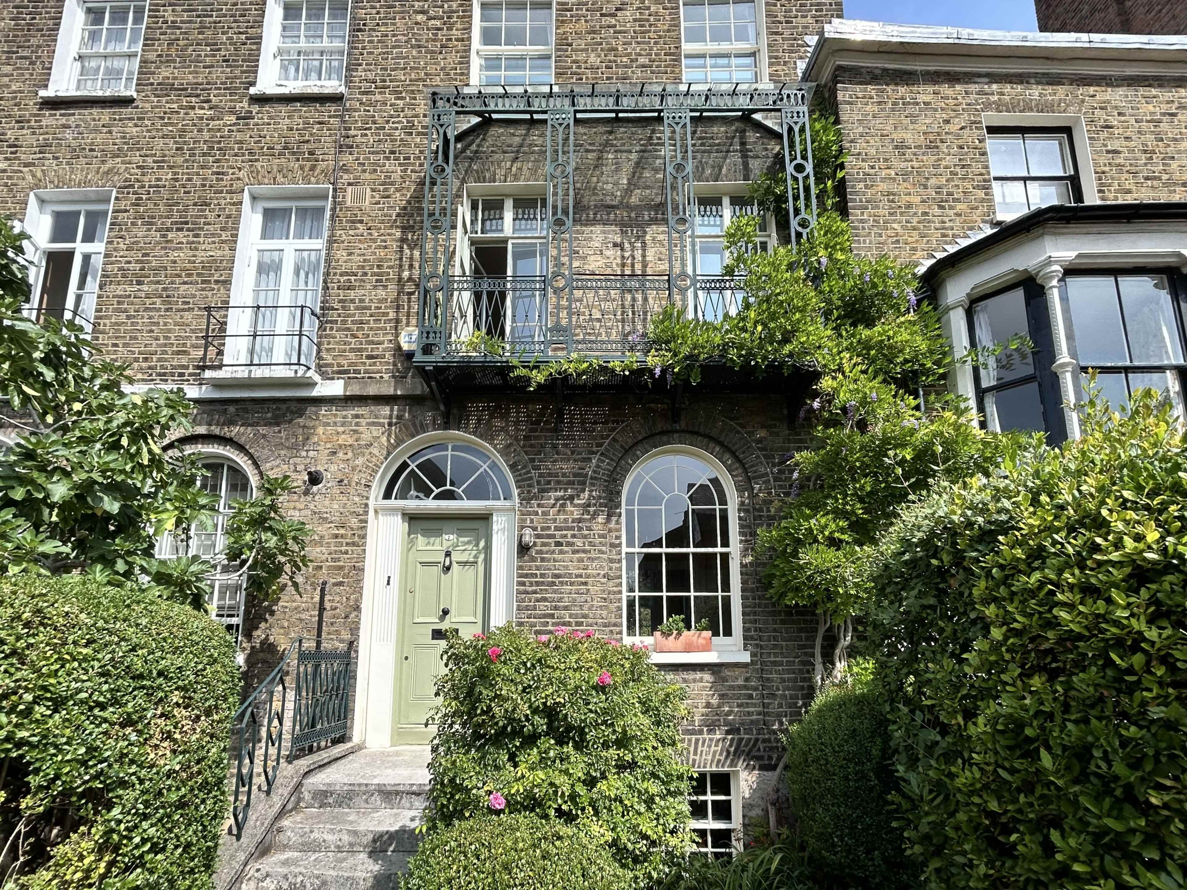 The image shows a brick townhouse with a green door and a balcony adorned with plants, surrounded by well-maintained hedges and bushes.