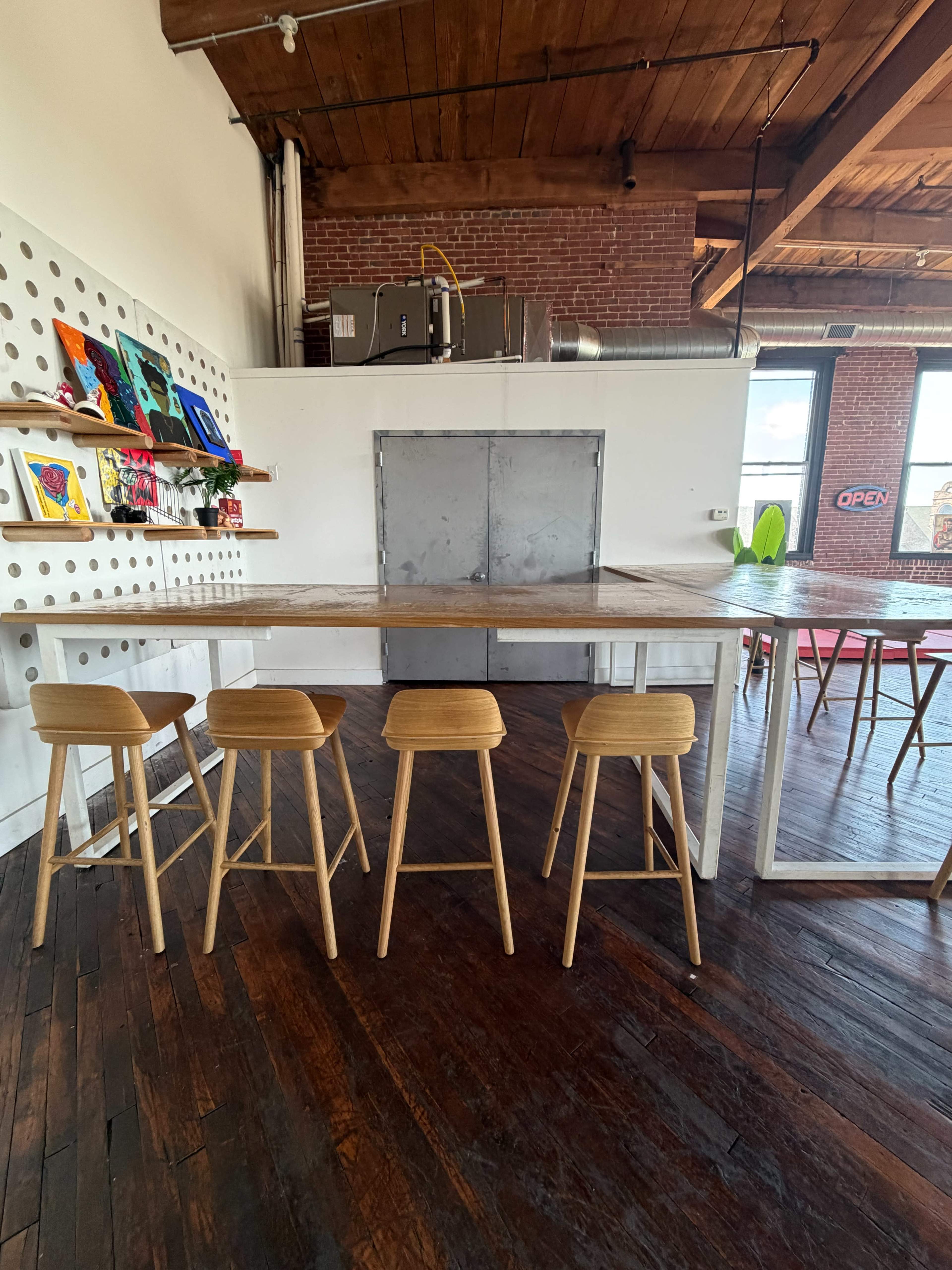 The image shows a modern workspace with a large wooden table surrounded by five wooden stools, featuring a backdrop of a textured wall and exposed brick.