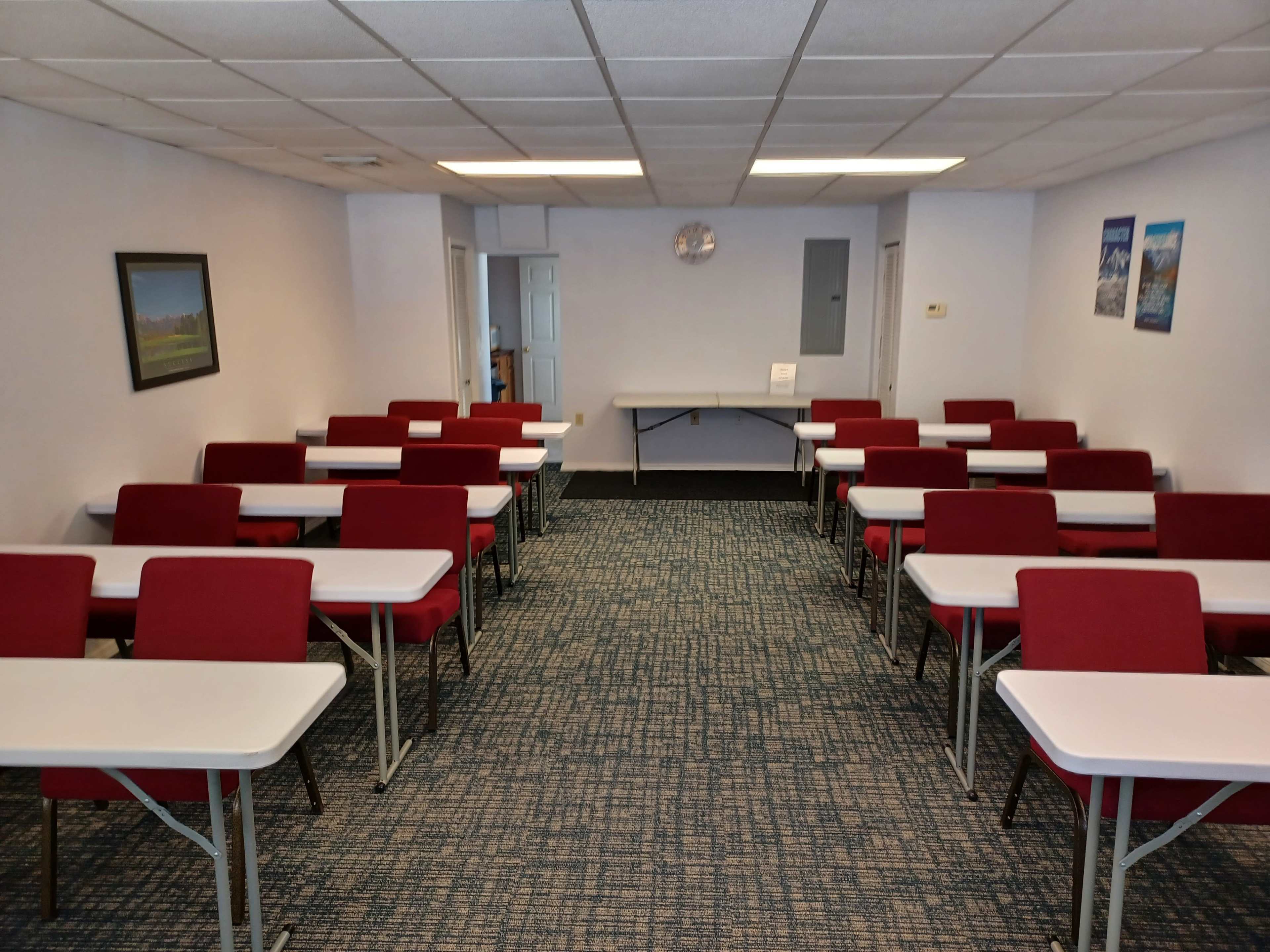 The image shows a classroom setting with rows of red chairs and tables arranged in a rectangular layout, against pale walls and a clock on the back wall.