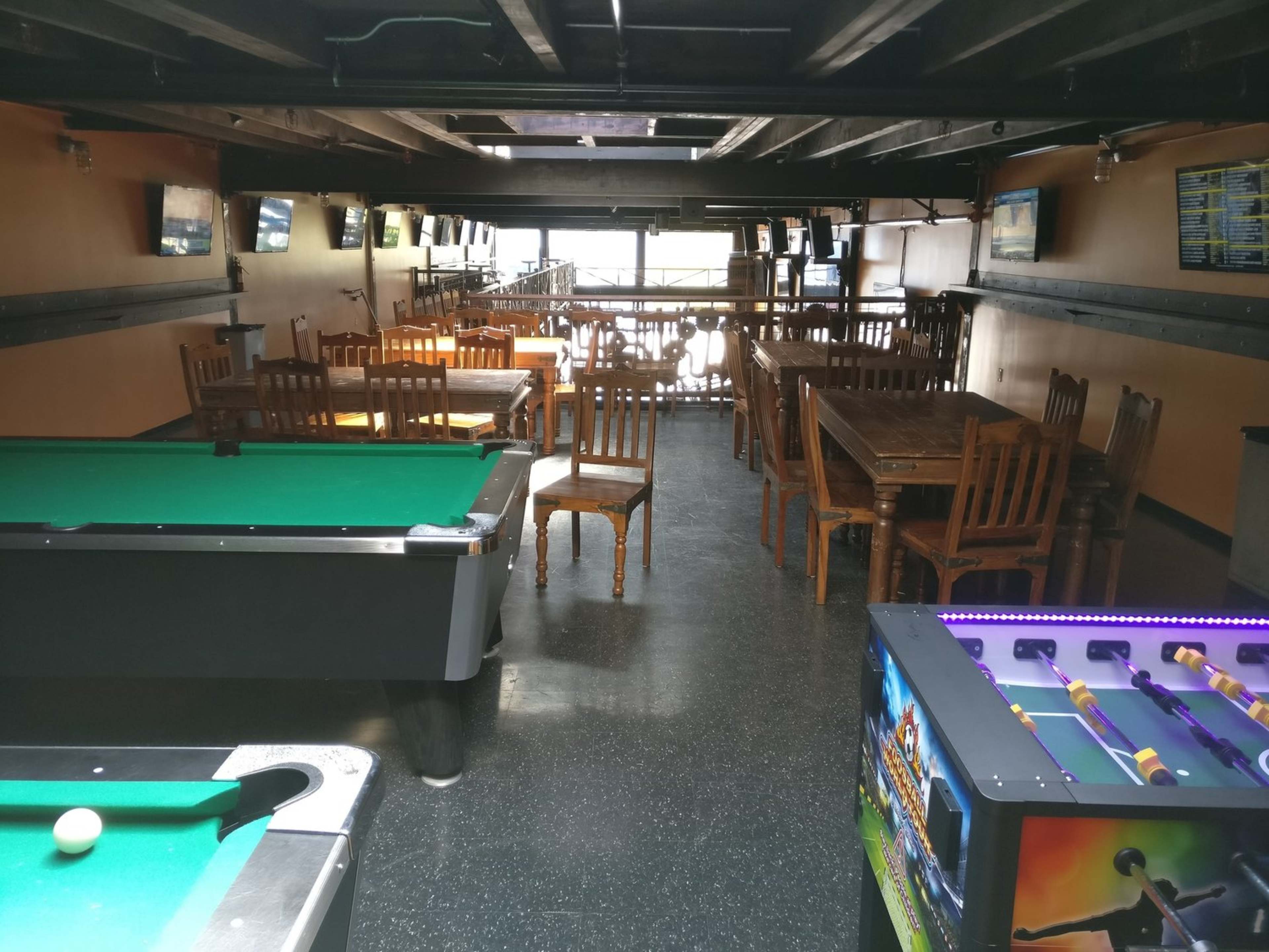 A spacious bar area featuring a pool table, foosball machine, and rows of wooden tables and chairs.