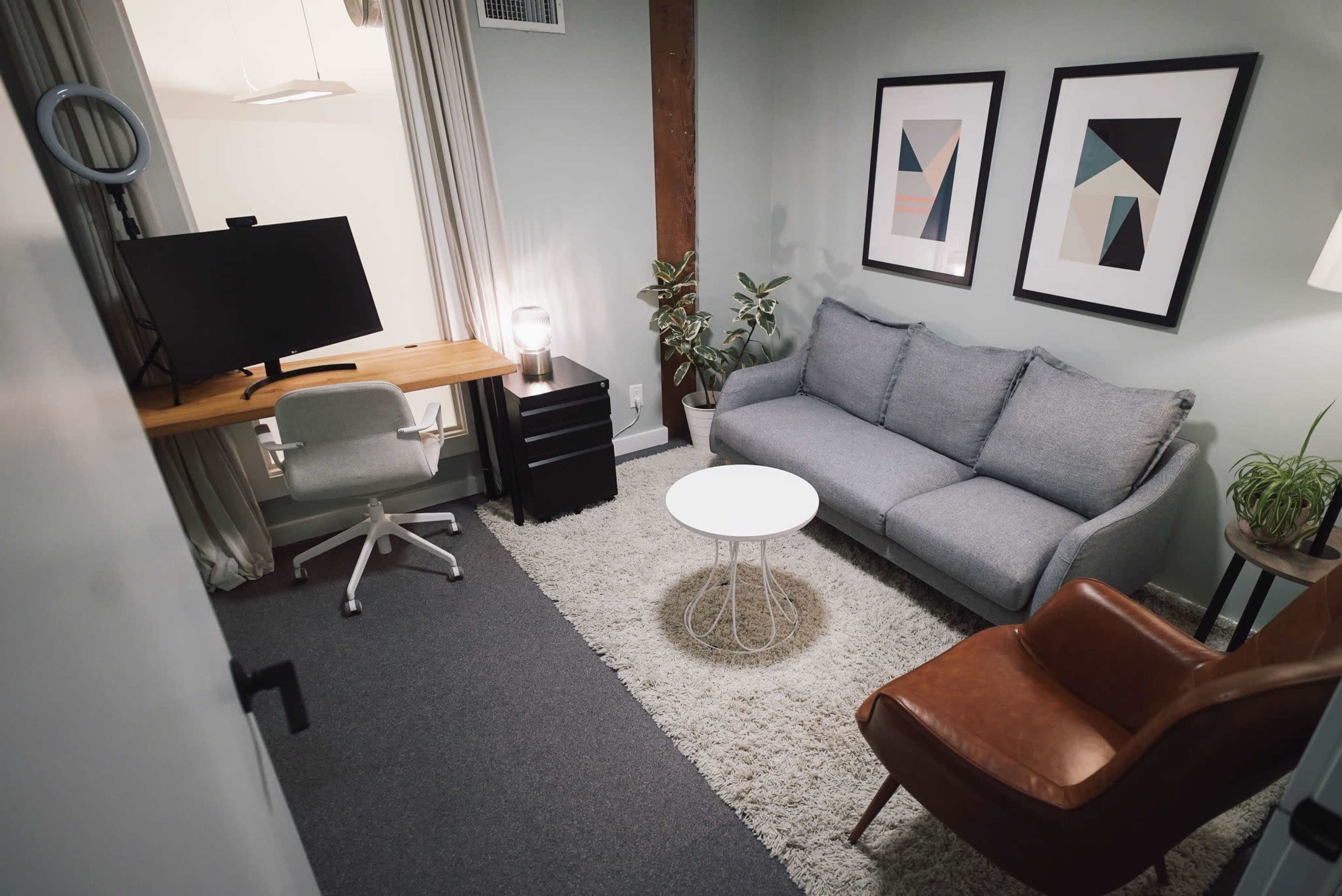 The image shows a cozy office space featuring a gray sofa, a small white table, an ergonomic chair at a wooden desk, and a brown armchair, all arranged around a soft area rug.