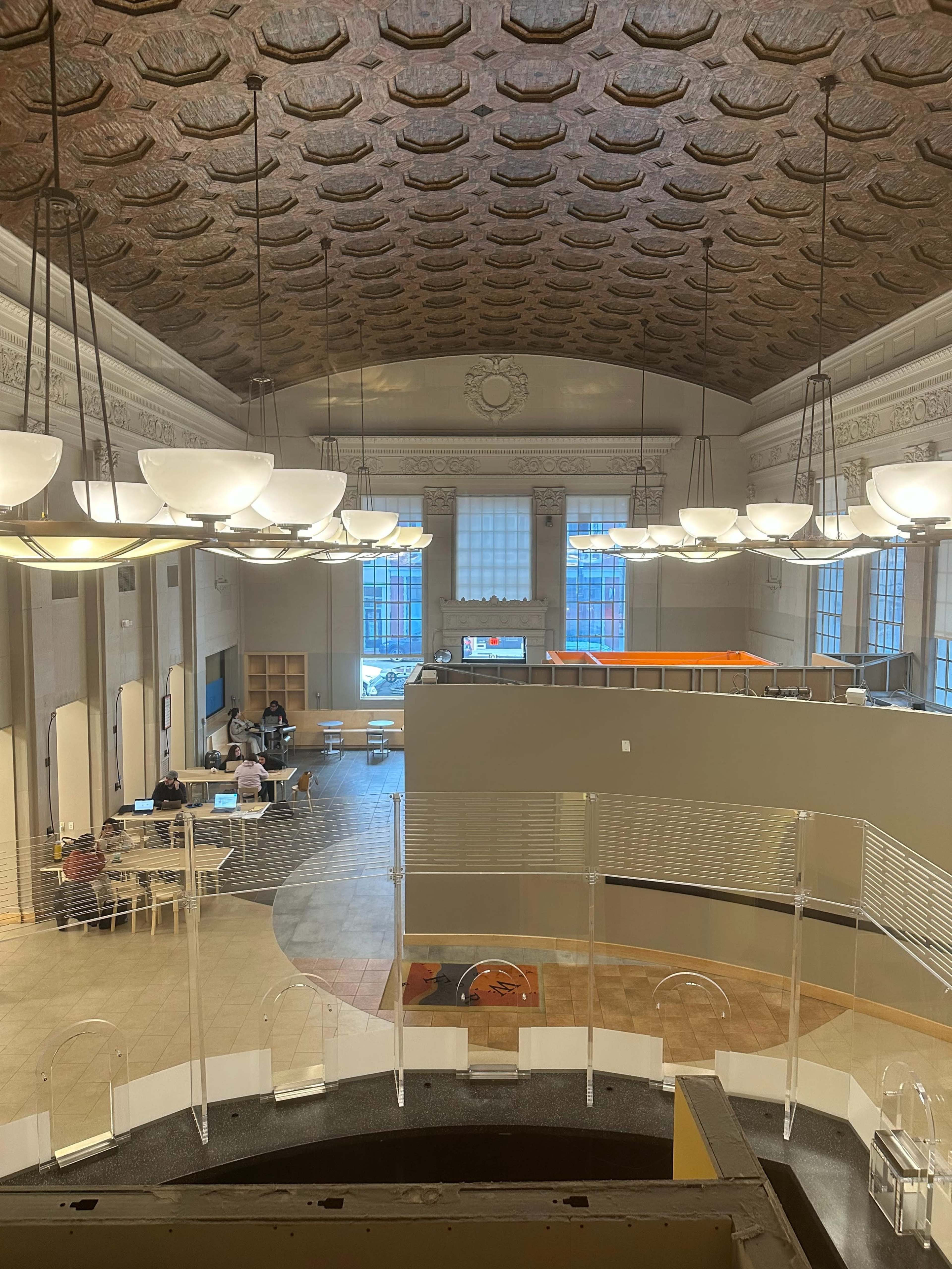 A spacious interior of a building with a high, decorative ceiling, large windows, and multiple tables where people are working.