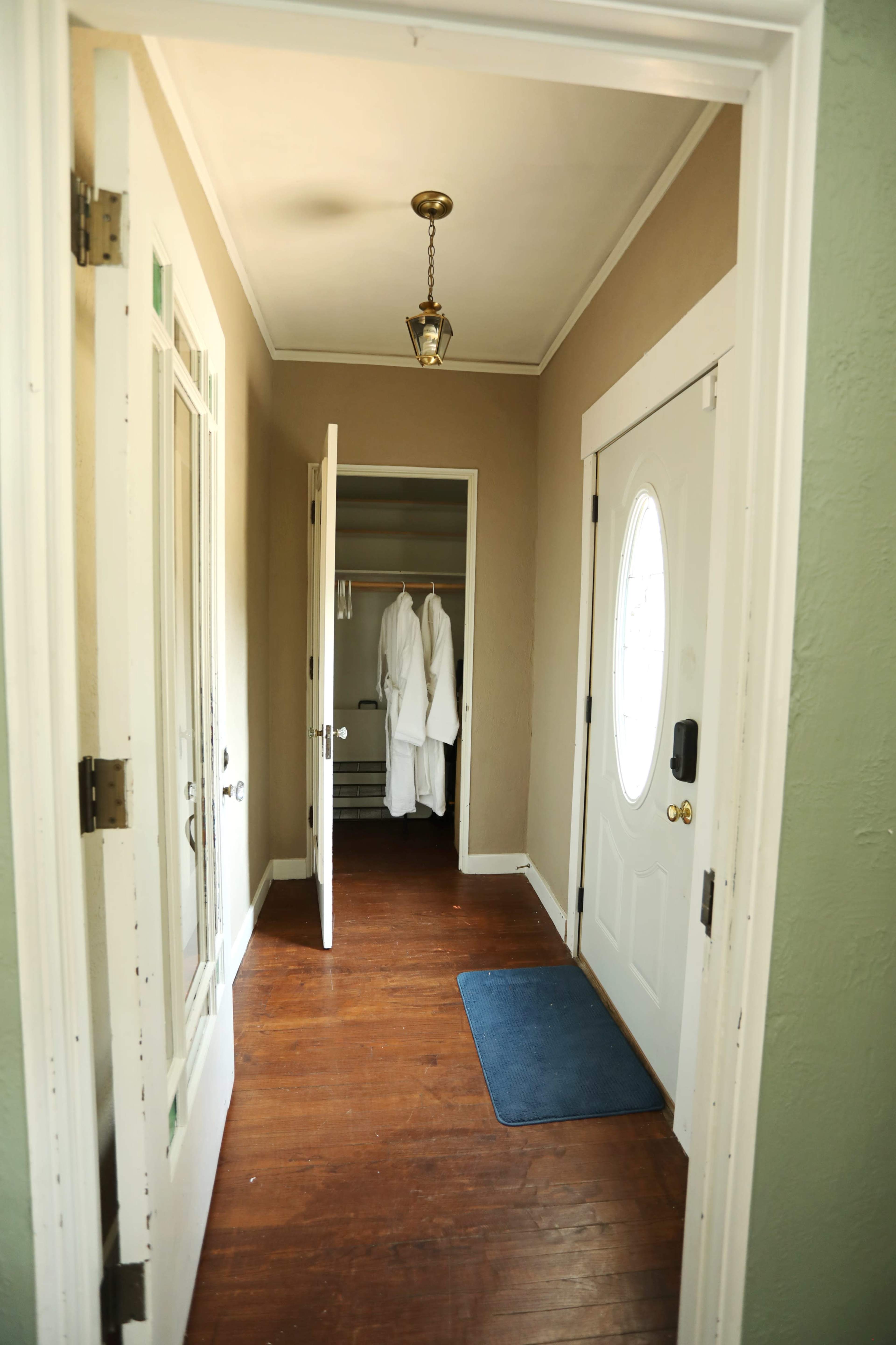 The image shows a hallway with a white door at one end, an open closet with two white robes hanging, and a blue mat on the wooden floor.
