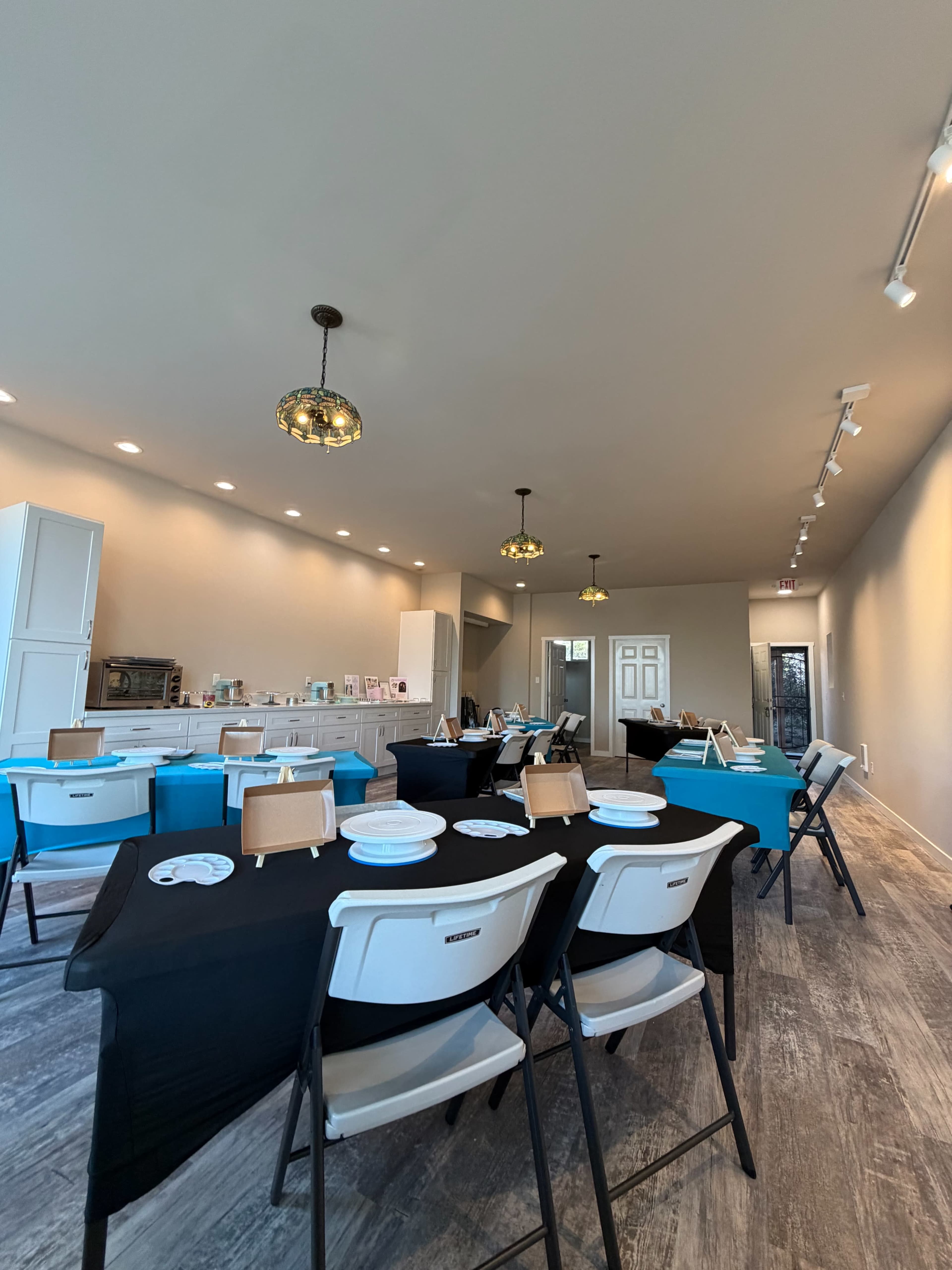 The image shows a spacious interior room set up for an event, featuring tables covered in black and turquoise tablecloths arranged with plates and easels.