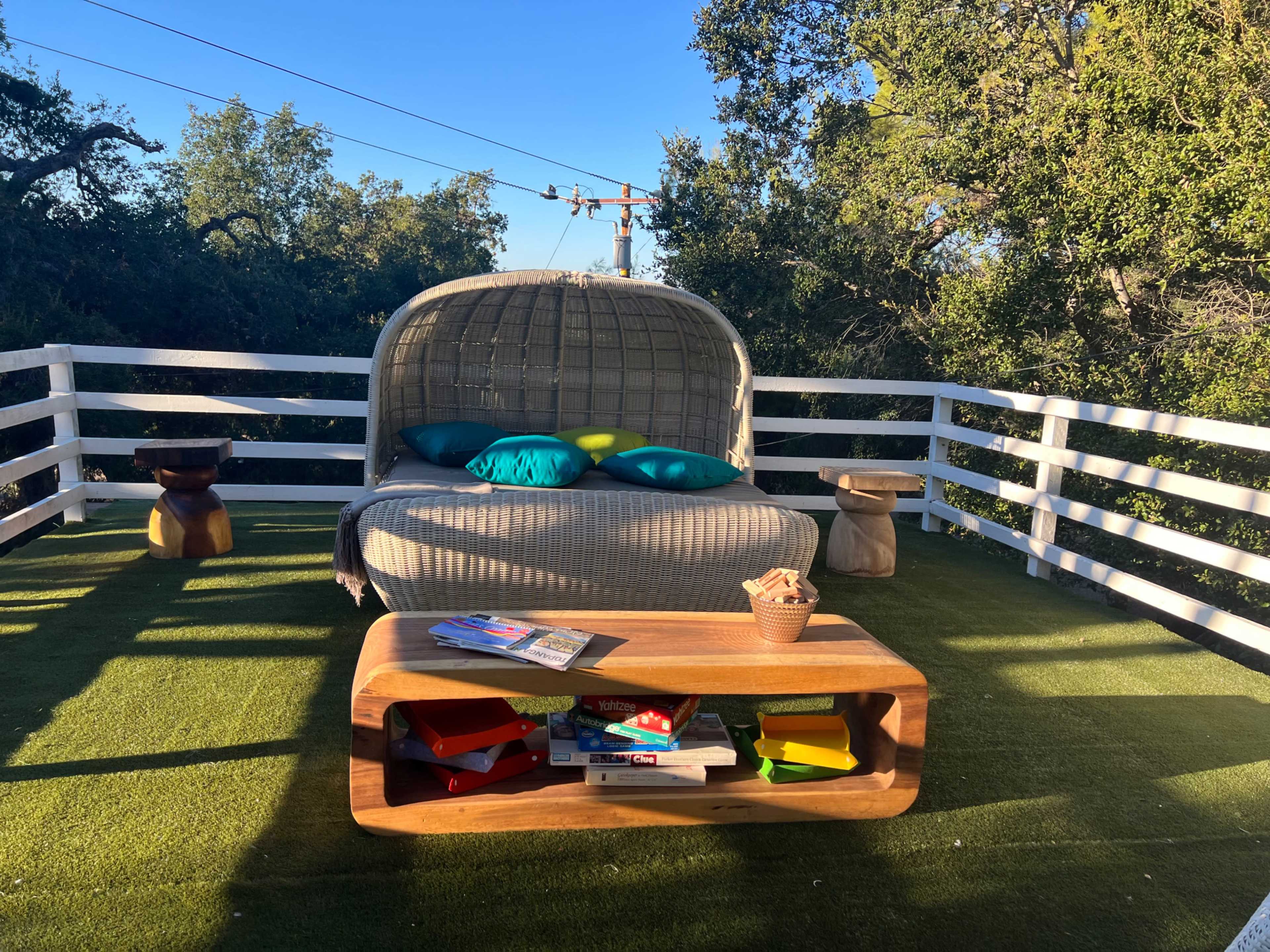 The scene features a cozy outdoor seating area with a large, cushioned lounge chair and a wooden coffee table, surrounded by greenery and a white fence.