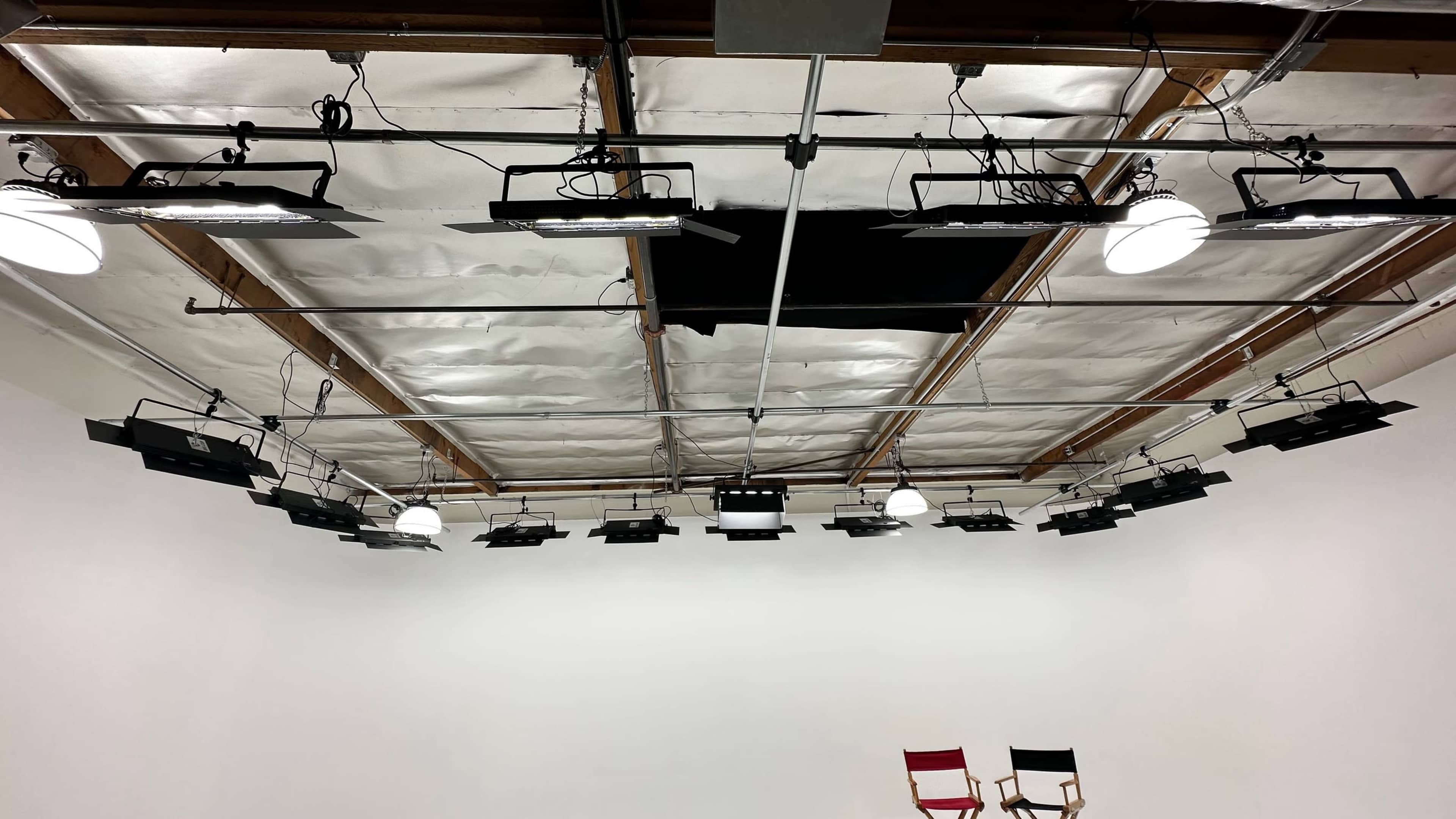 A well-lit studio with a white backdrop and multiple overhead lights mounted on a wooden ceiling.