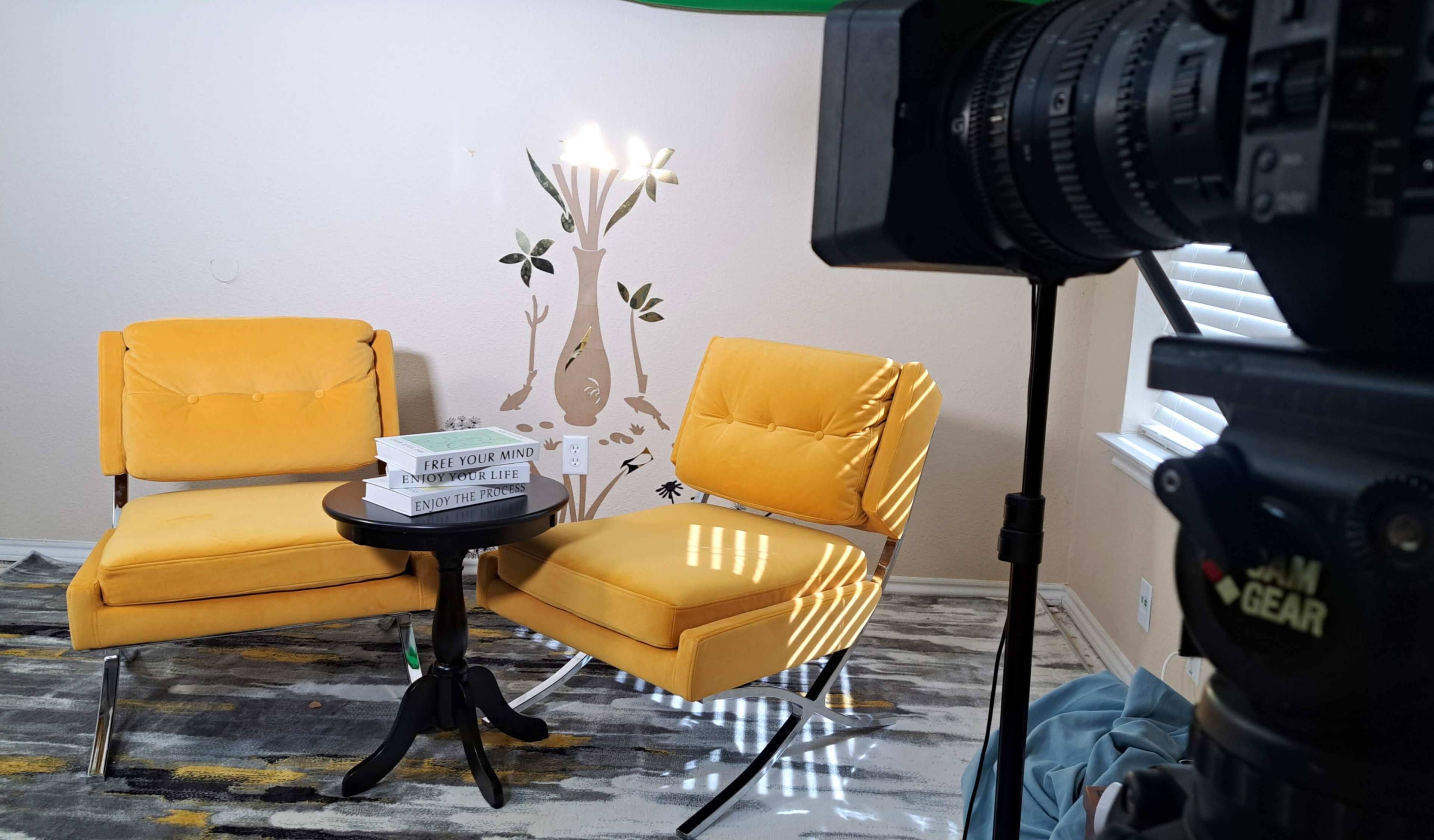 The image shows a cozy interior scene featuring two yellow chairs, a small black table with books, and a decorative wall mural of a tree.