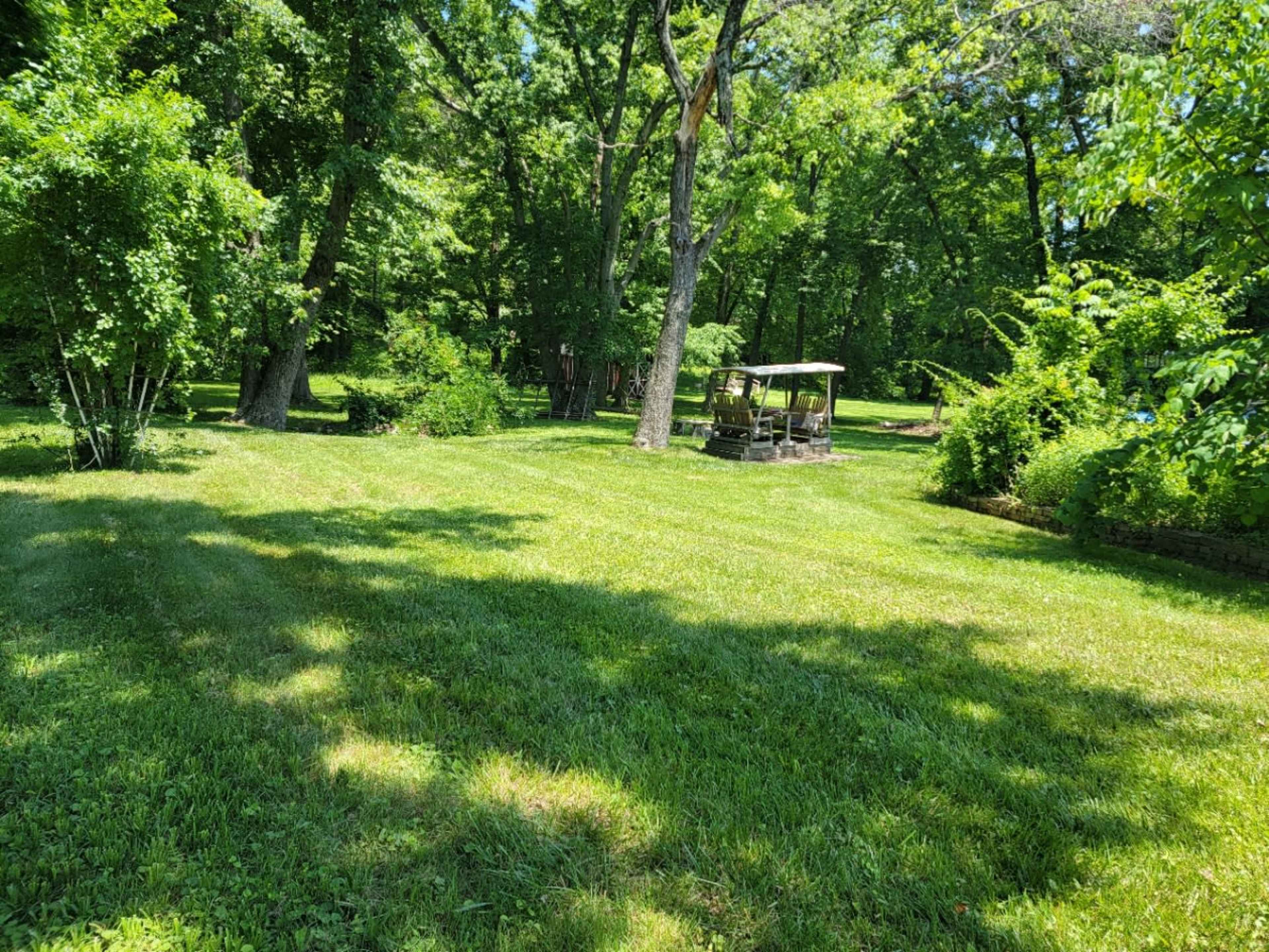 A grassy area in a wooded park features a small gazebo surrounded by lush green trees.