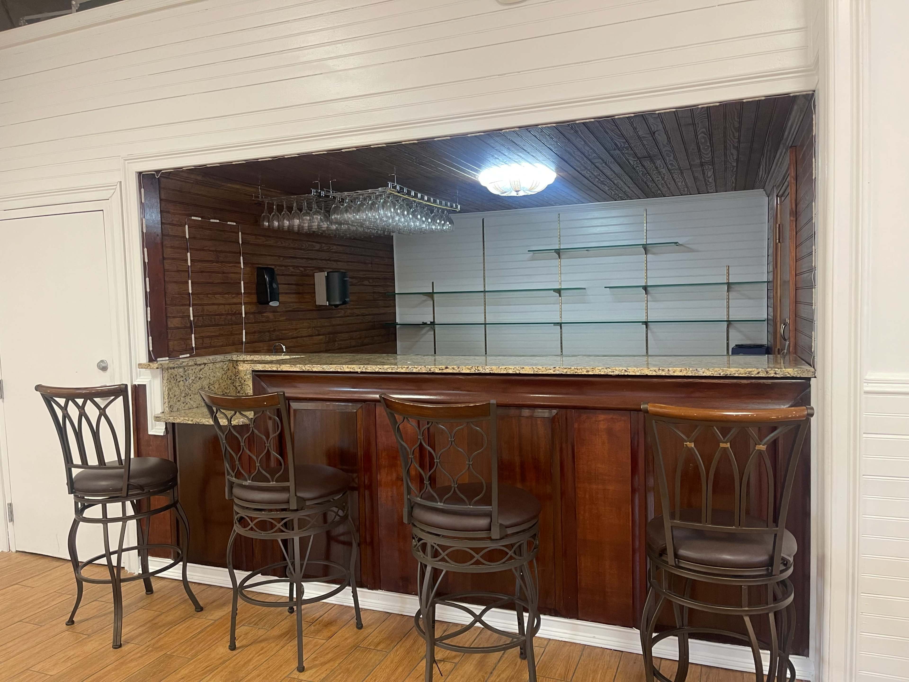 The image shows a bar area with a granite countertop and four metal stools, situated against a wooden-paneled wall, with empty shelves in the background.