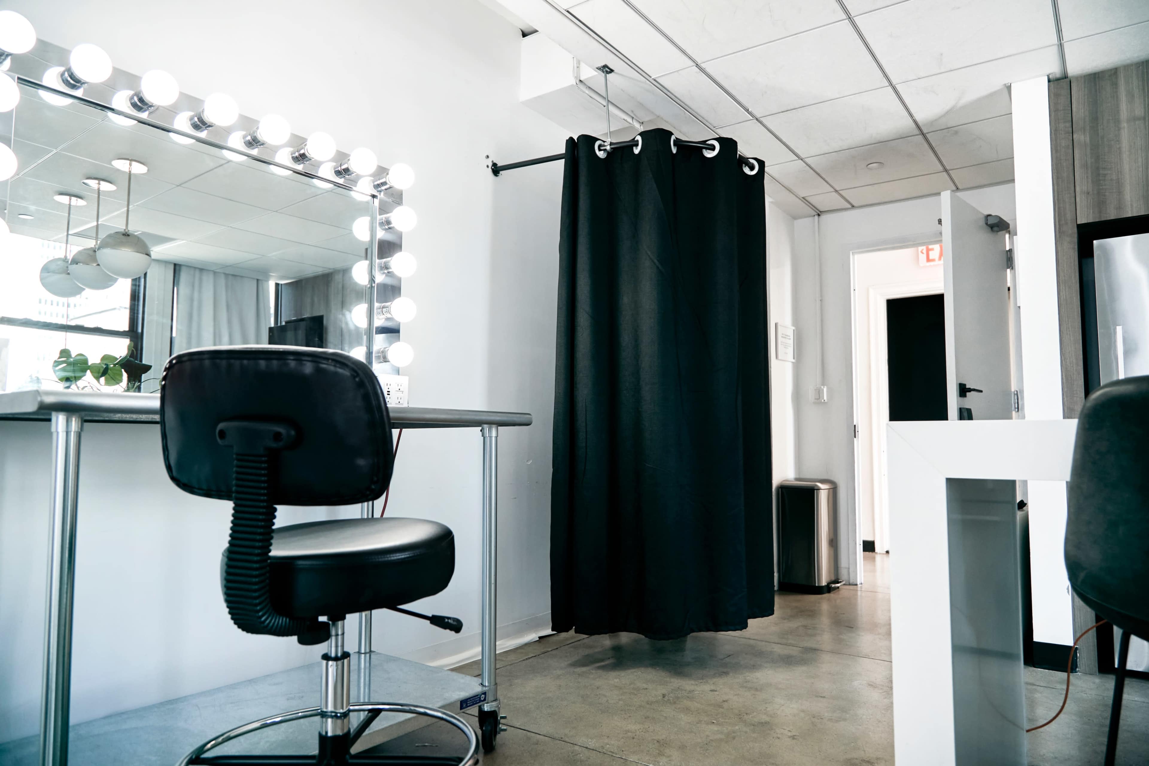 A modern beauty salon interior featuring a vanity mirror with lights, a straight-backed chair, a black curtain, and a doorway at the far end.