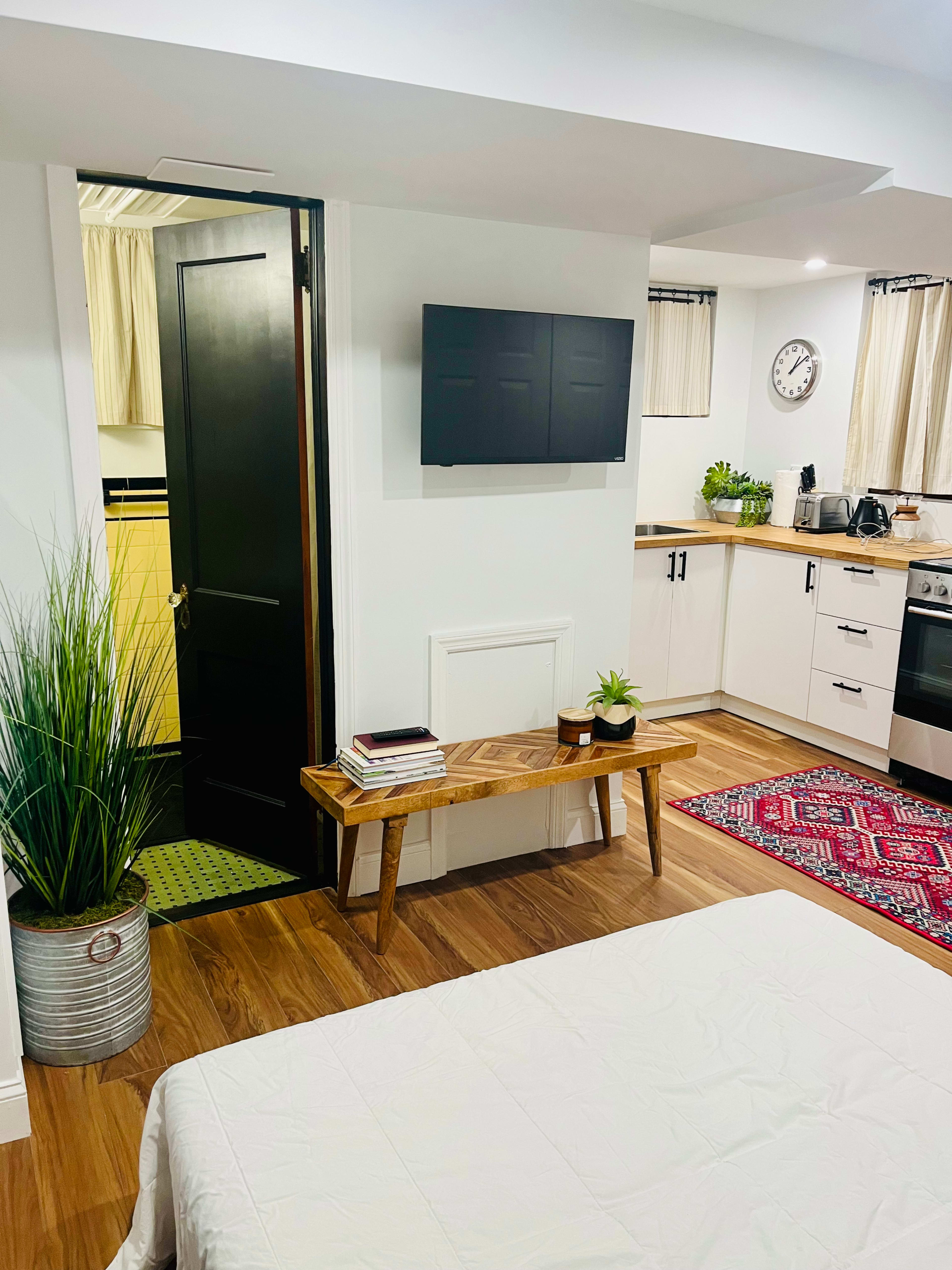 The image shows a small studio apartment with a kitchenette, a bathroom door, a wooden bench, and a bed in the foreground.