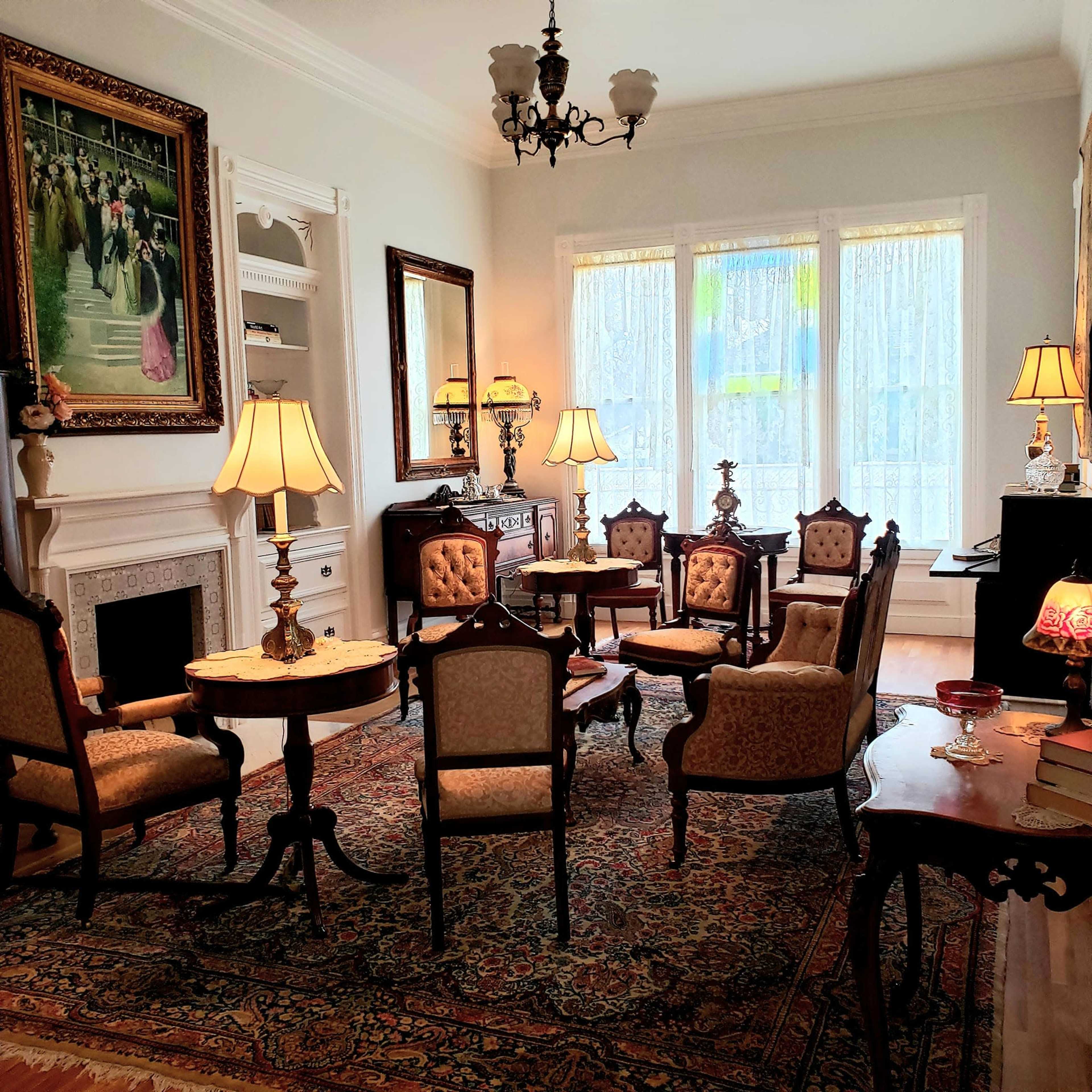 The room features elegantly furnished antique chairs and tables, with a large painting on the wall and natural light streaming through stained glass windows.