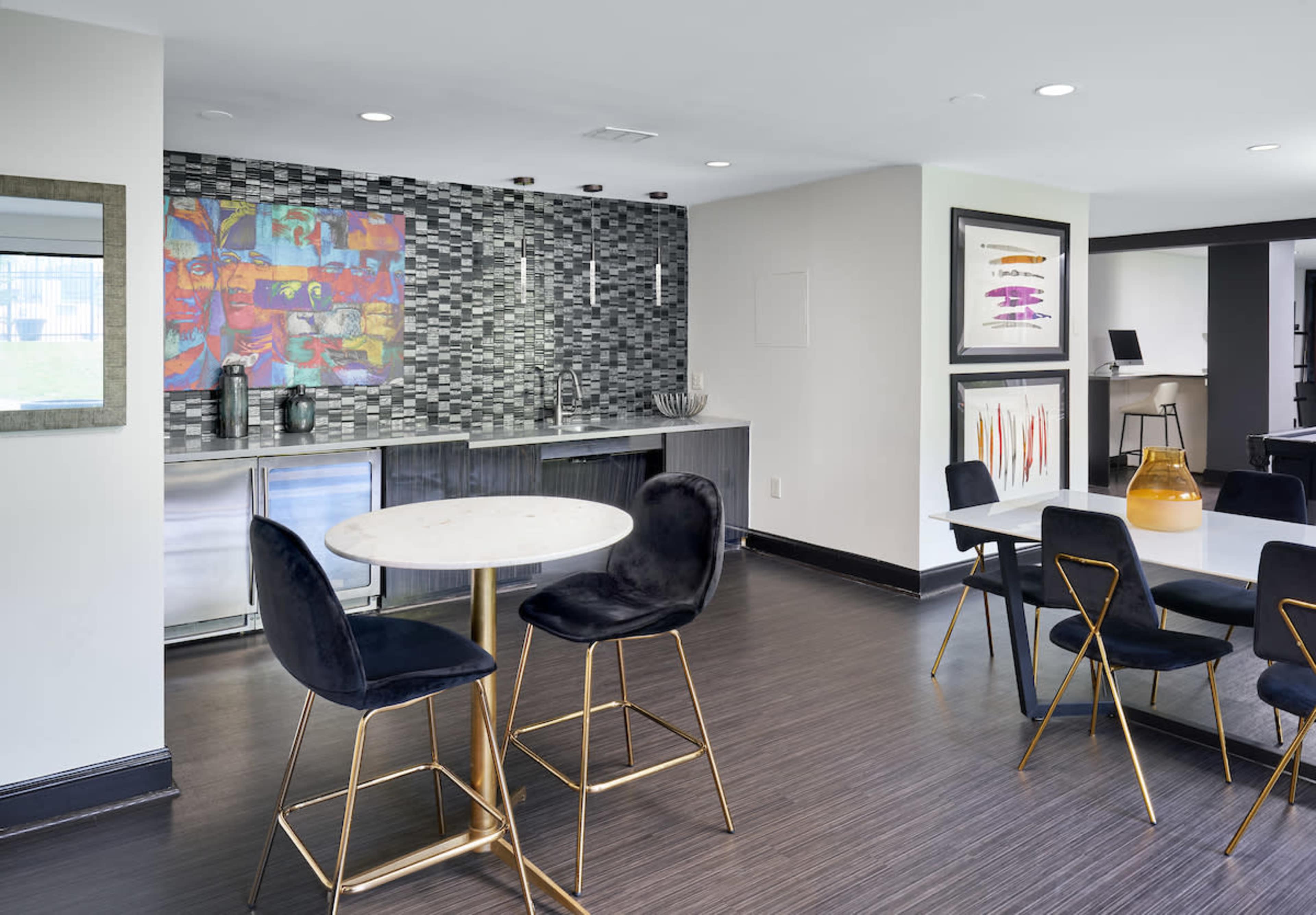 A modern lounge area featuring a marble-top table with two black chairs, a kitchenette with a tiled backsplash, and various artworks displayed on the walls.