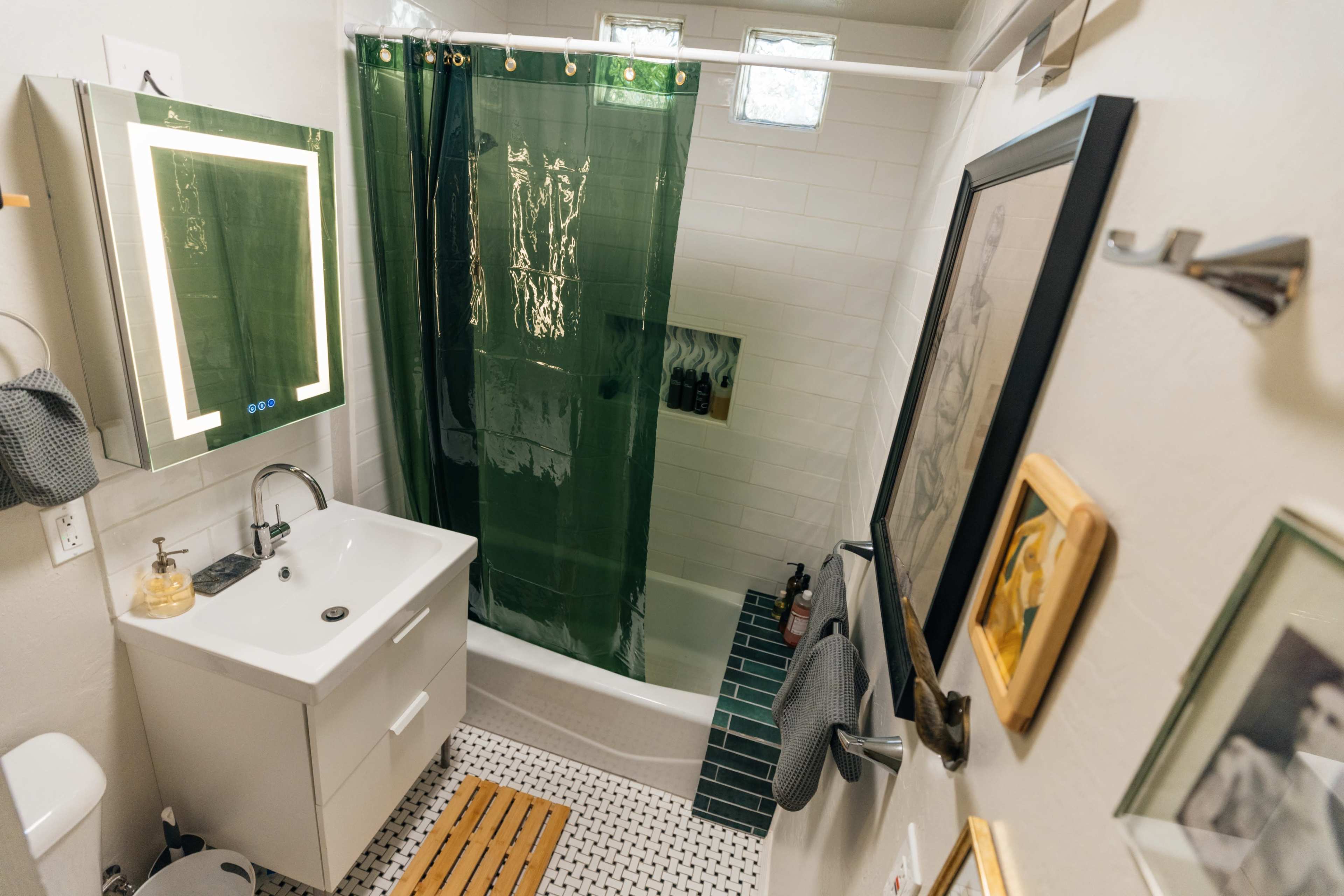 A bathroom features a shower with a green curtain, a white sink with a cabinet, and patterned black and white floor tiles.