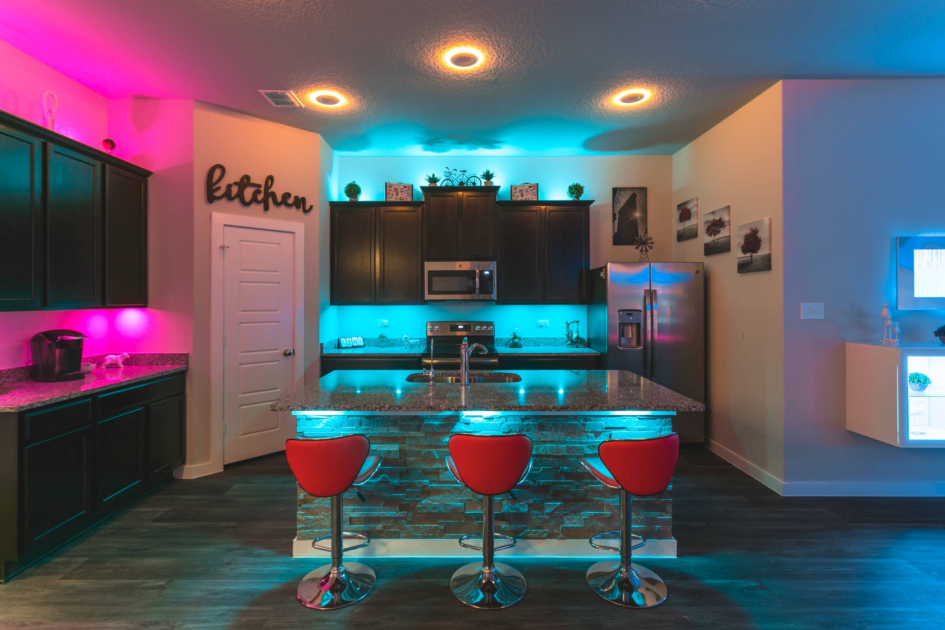 A modern kitchen features dark cabinetry, a granite countertop, and illuminated shelving with blue and pink lighting.