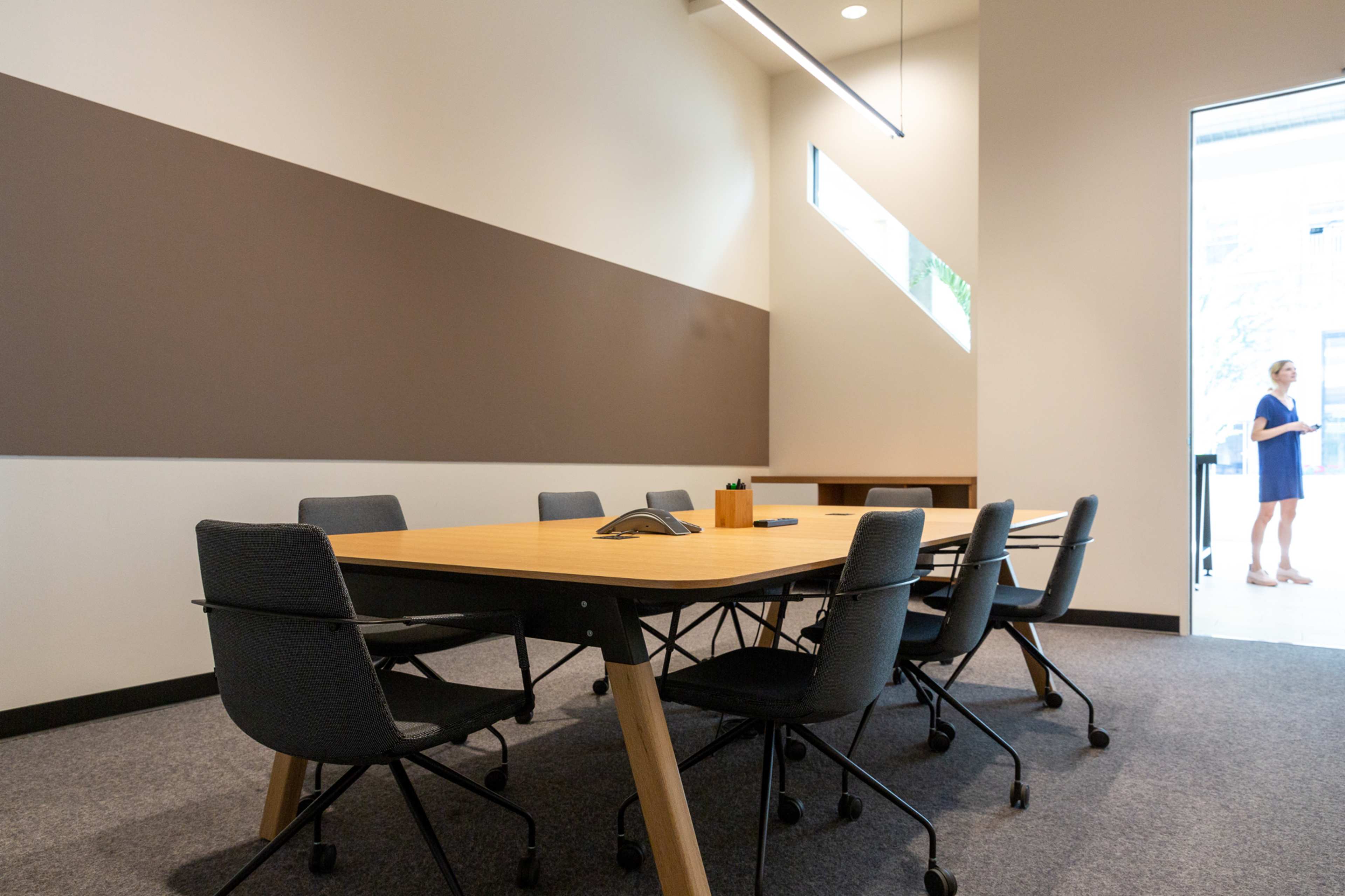 A conference room features a large wooden table surrounded by rolling chairs, with a doorway leading to an adjacent space.