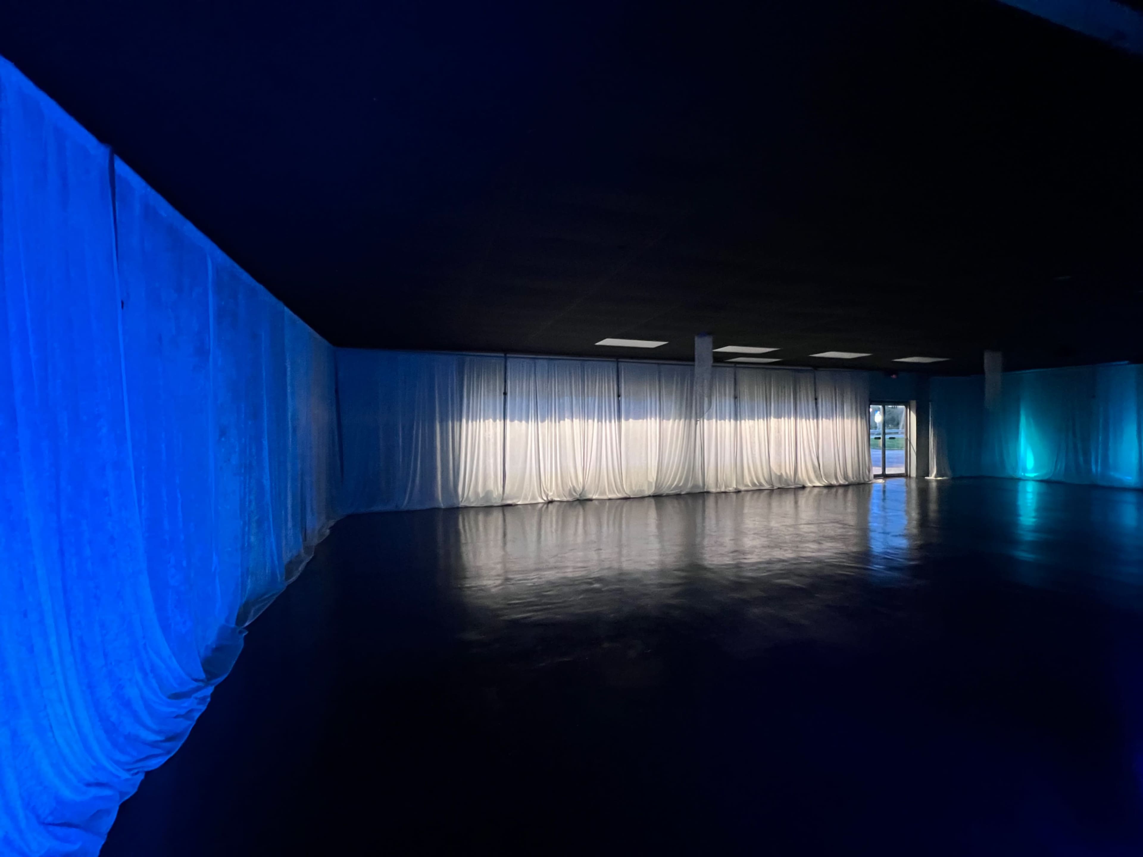 An empty room with dark flooring and blue-lit sheer curtains lining the walls.