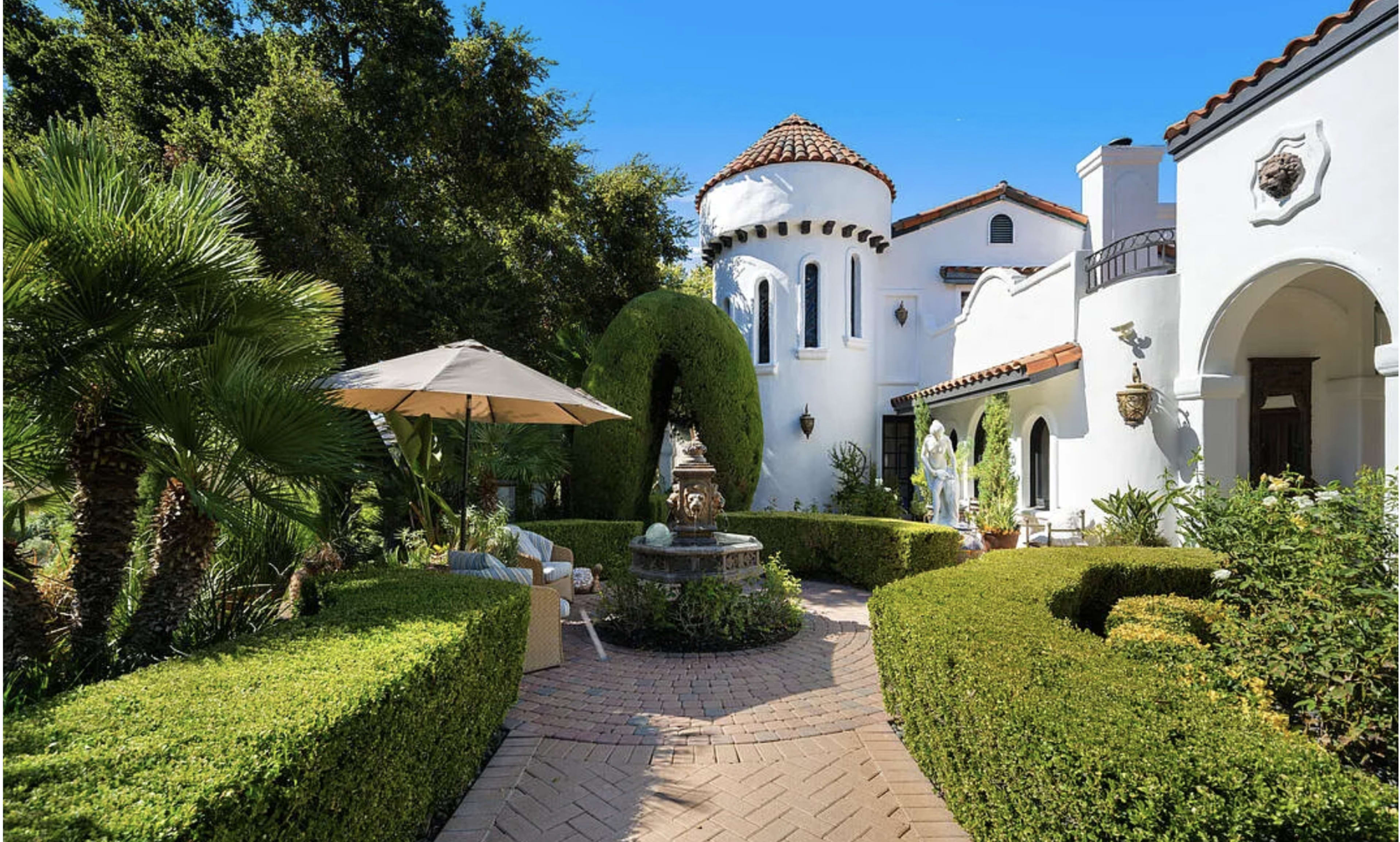 The image features a landscaped courtyard with a fountain surrounded by hedges, palm trees, and a white stucco building with a tiled rooftop tower.