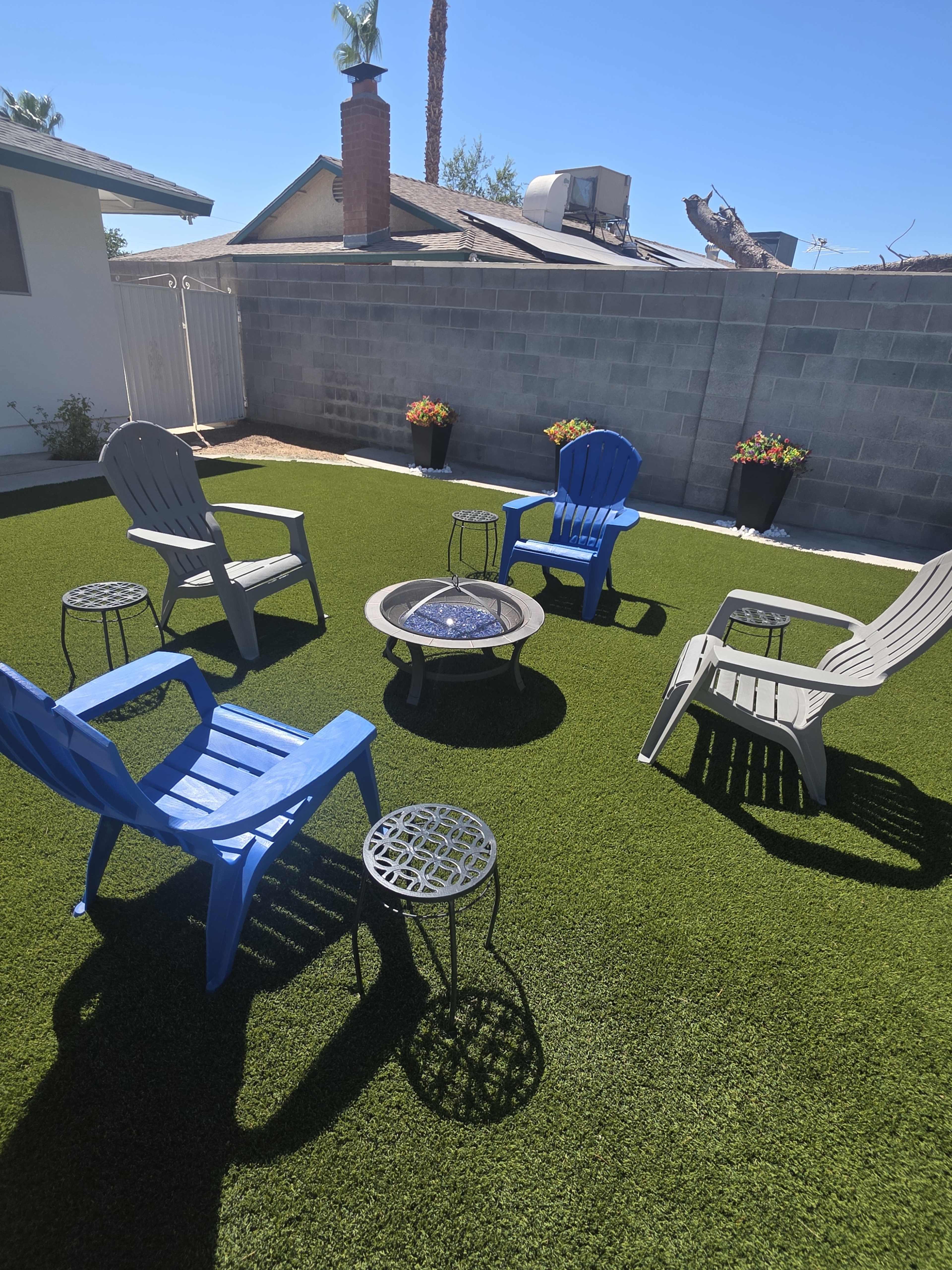 A small outdoor area features a circular arrangement of six chairs and a fire pit on synthetic grass, surrounded by a gray wall and potted plants.
