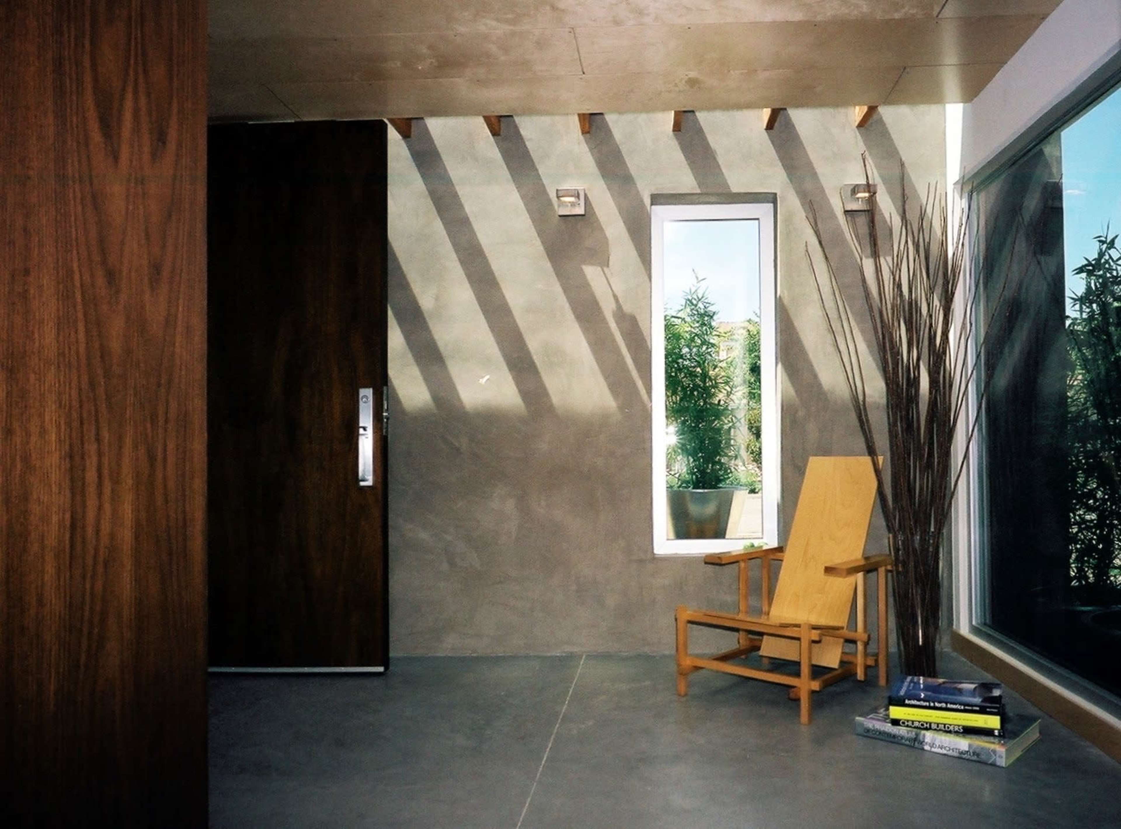 A modern interior space featuring a wooden chair, a large window, and a solid door against a concrete wall with natural light casting shadows.