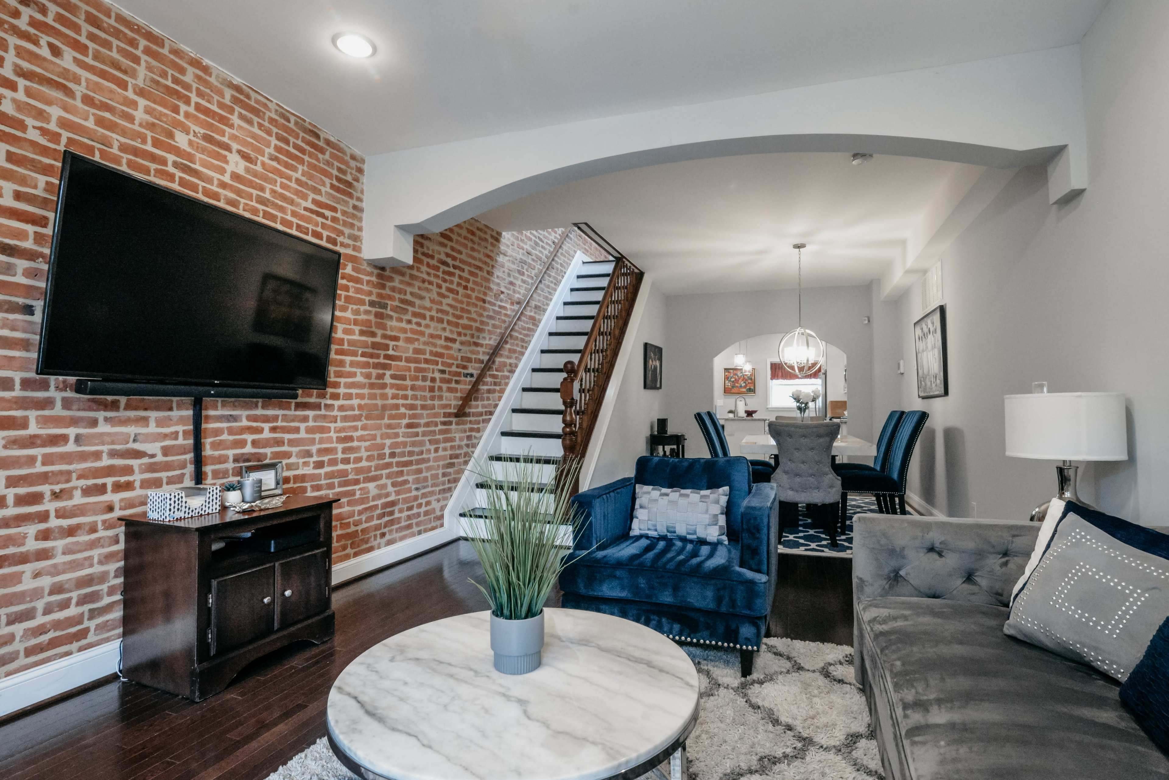 A modern living room with a brick wall, a large TV mounted on the wall, and a seating area featuring a blue sofa and a round coffee table.