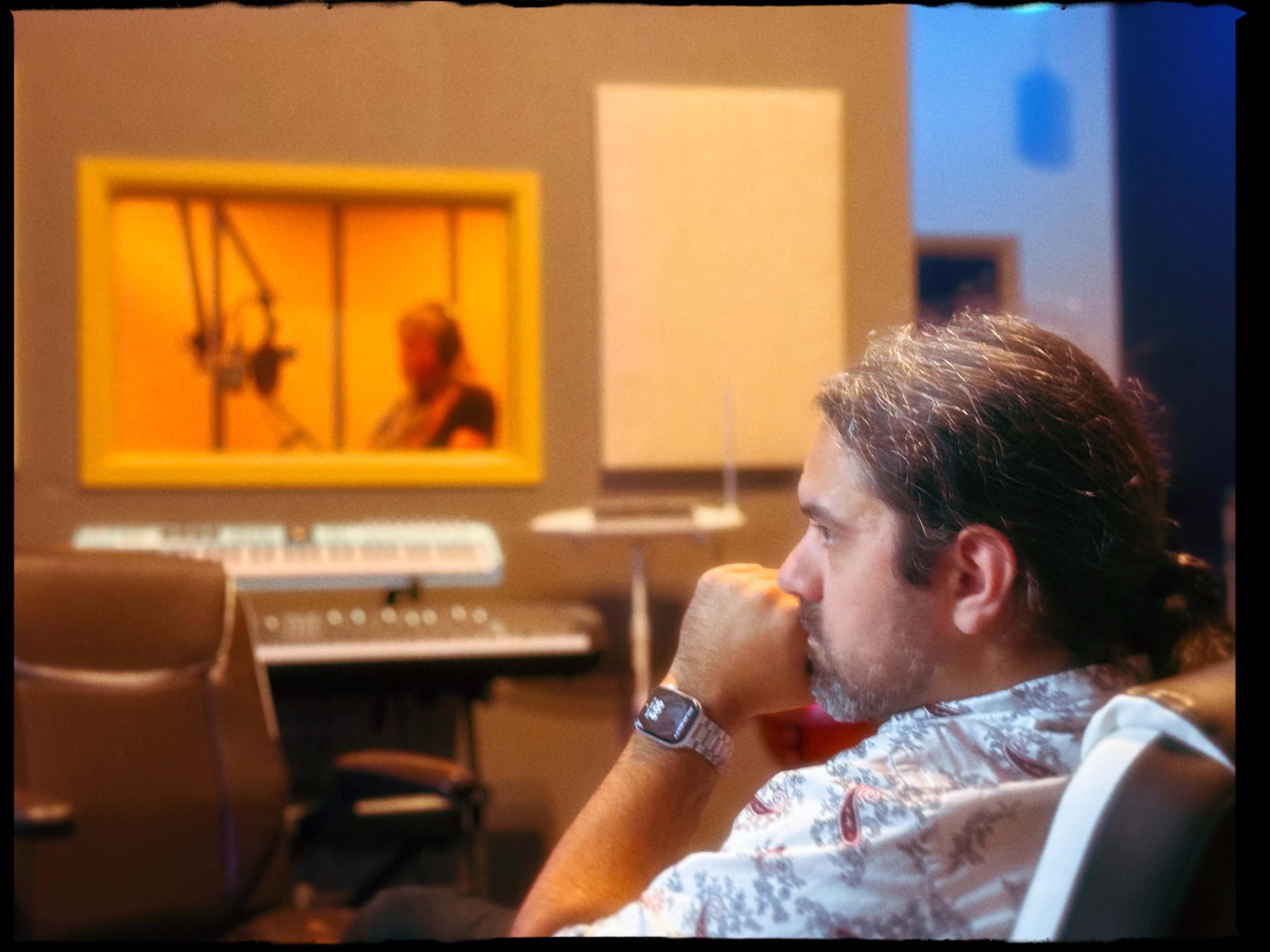A man sits in a recording studio, observing another person through a window as they record music.