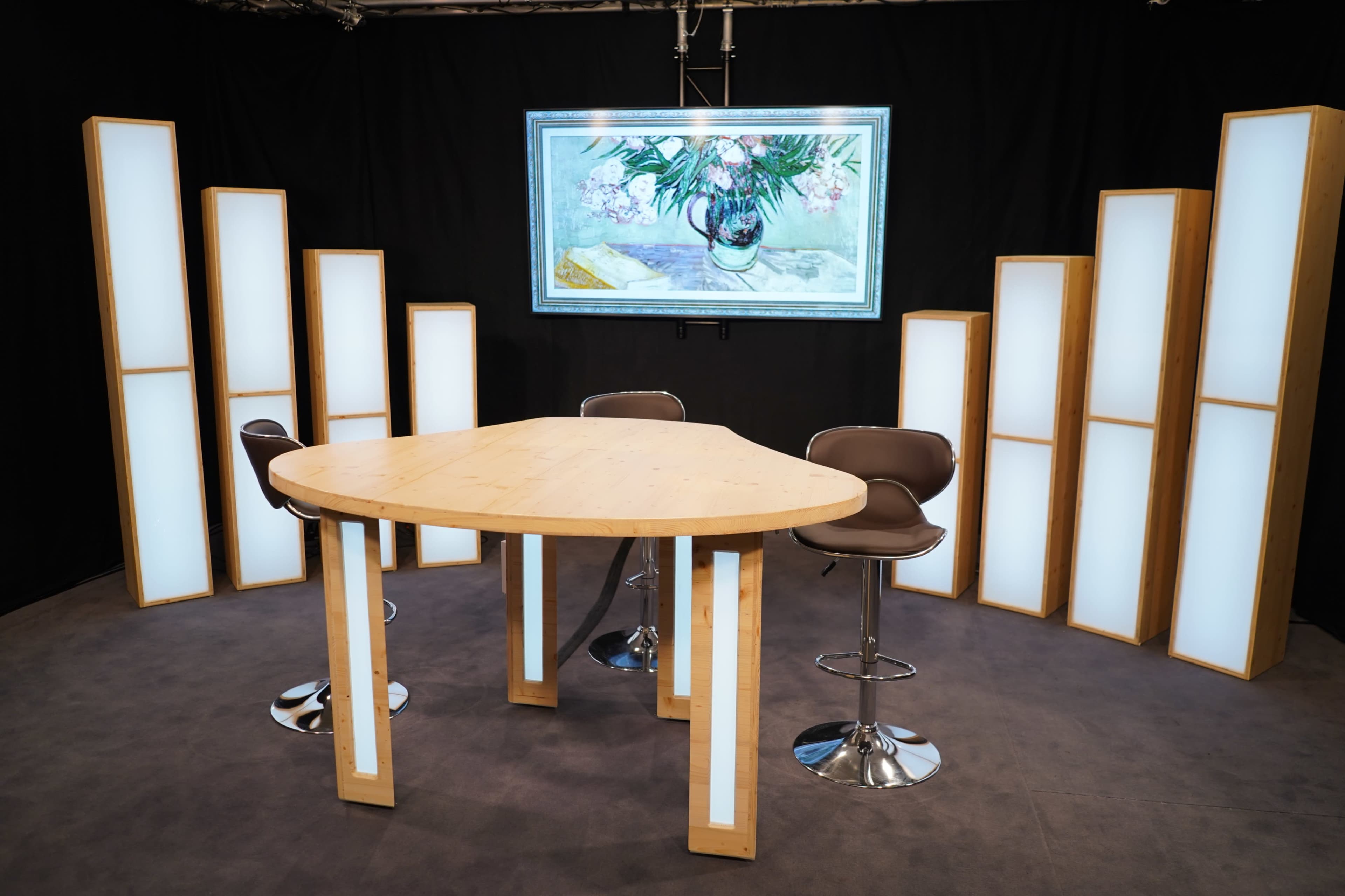 A modern studio setup features a triangular wooden table surrounded by high stools, illuminated by tall, backlit panels and a large artwork displayed above.