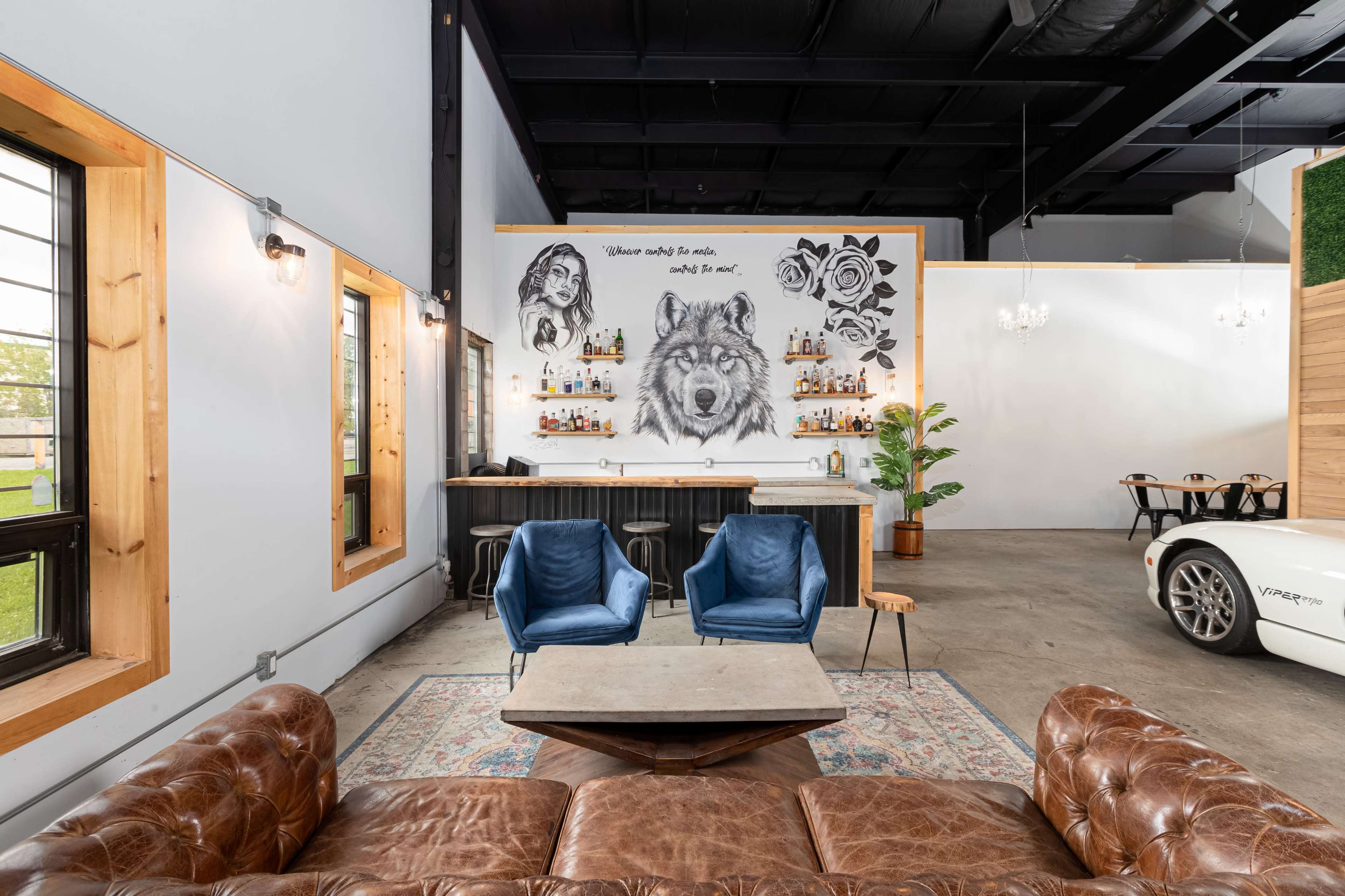 A spacious interior featuring a brown leather sofa, blue armchairs, a wooden bar with a wall mural of a wolf and decorative items, along with a dining area in the background.