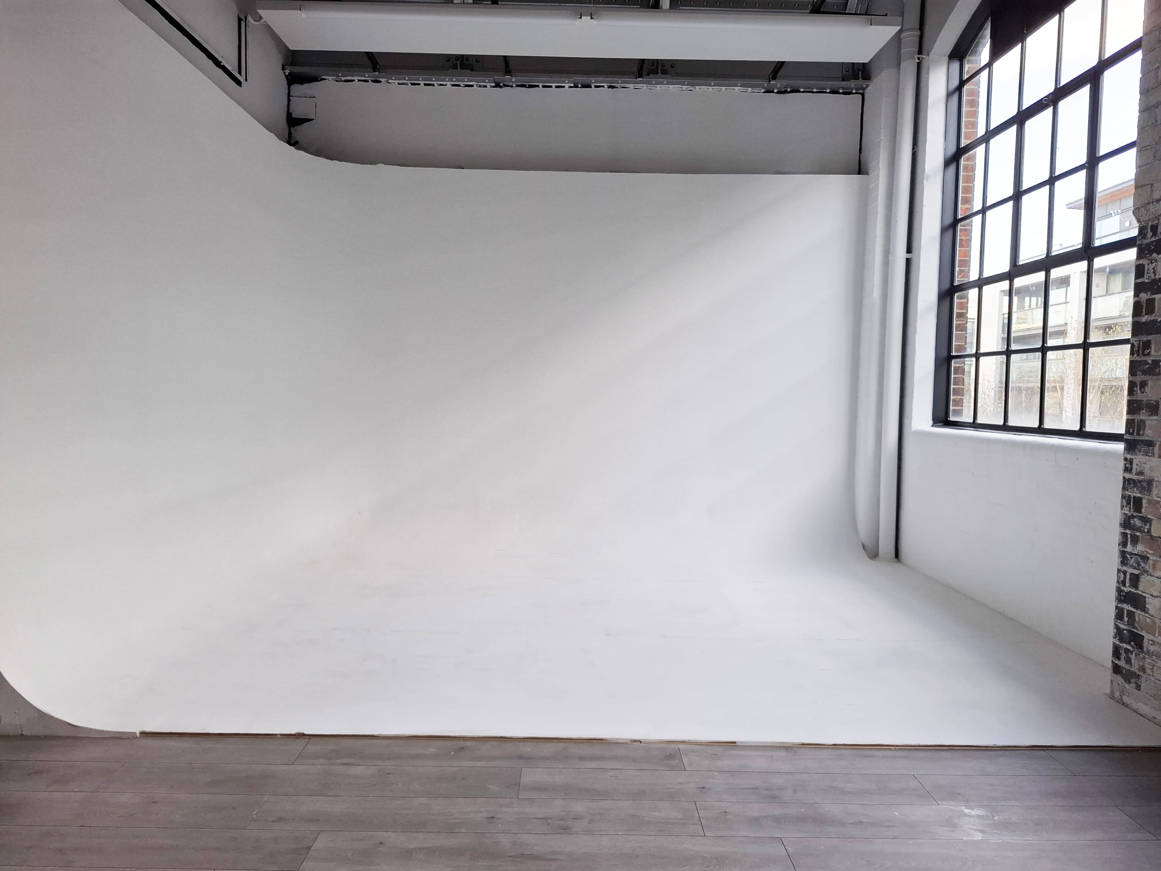 The image shows a spacious, empty photo studio with a white seamless backdrop and large windows allowing natural light to enter.