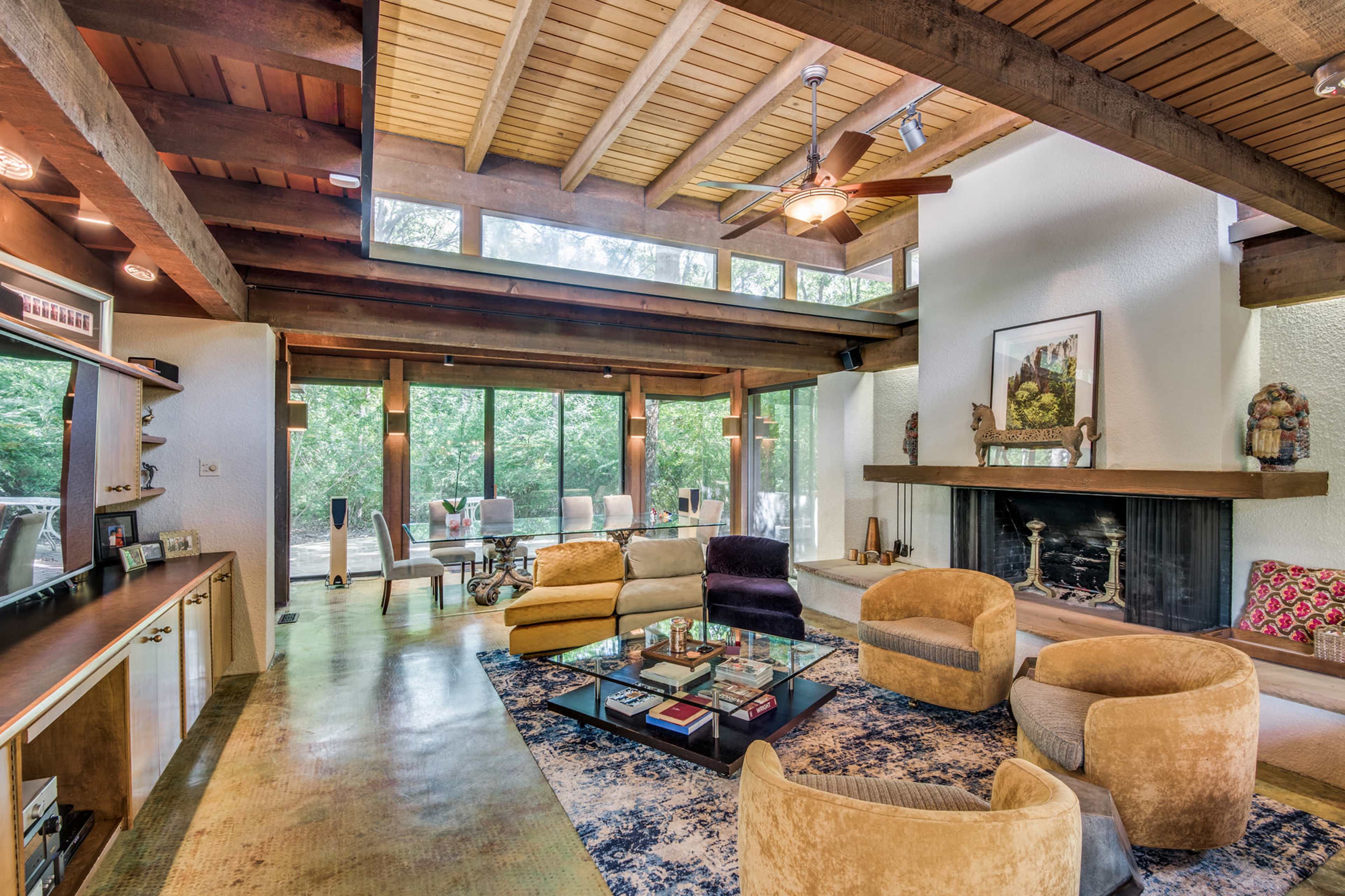 A modern living room with wooden beams, a stone fireplace, and large windows that overlook a wooded area.
