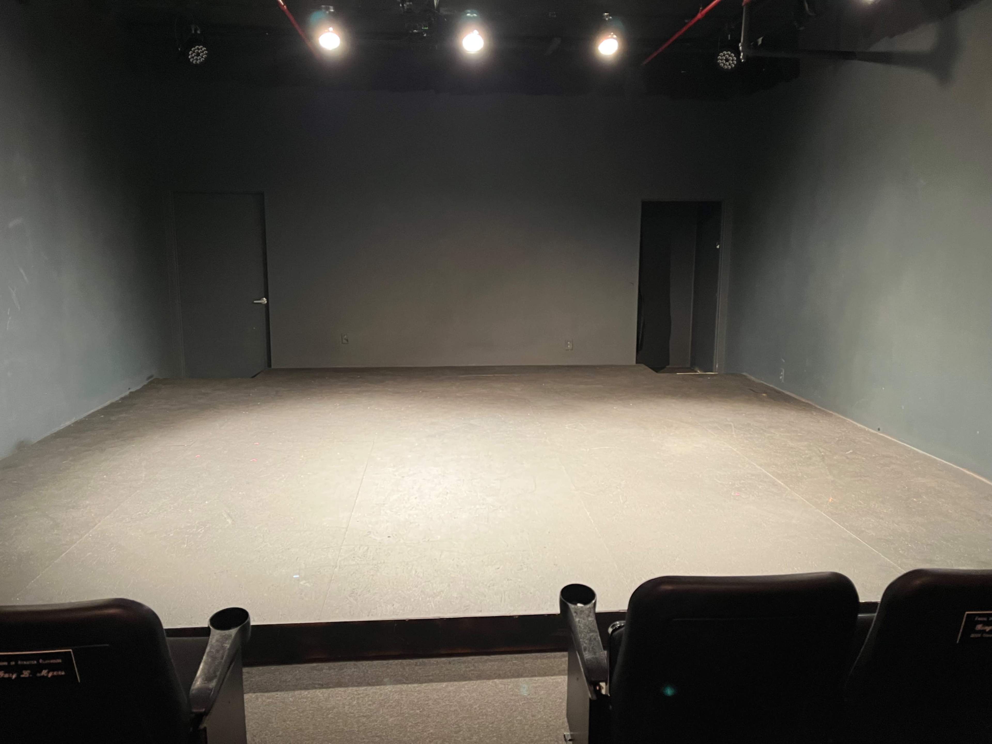 The image shows a sparsely furnished theater stage with two doors and overhead lights, facing a row of empty black seats.