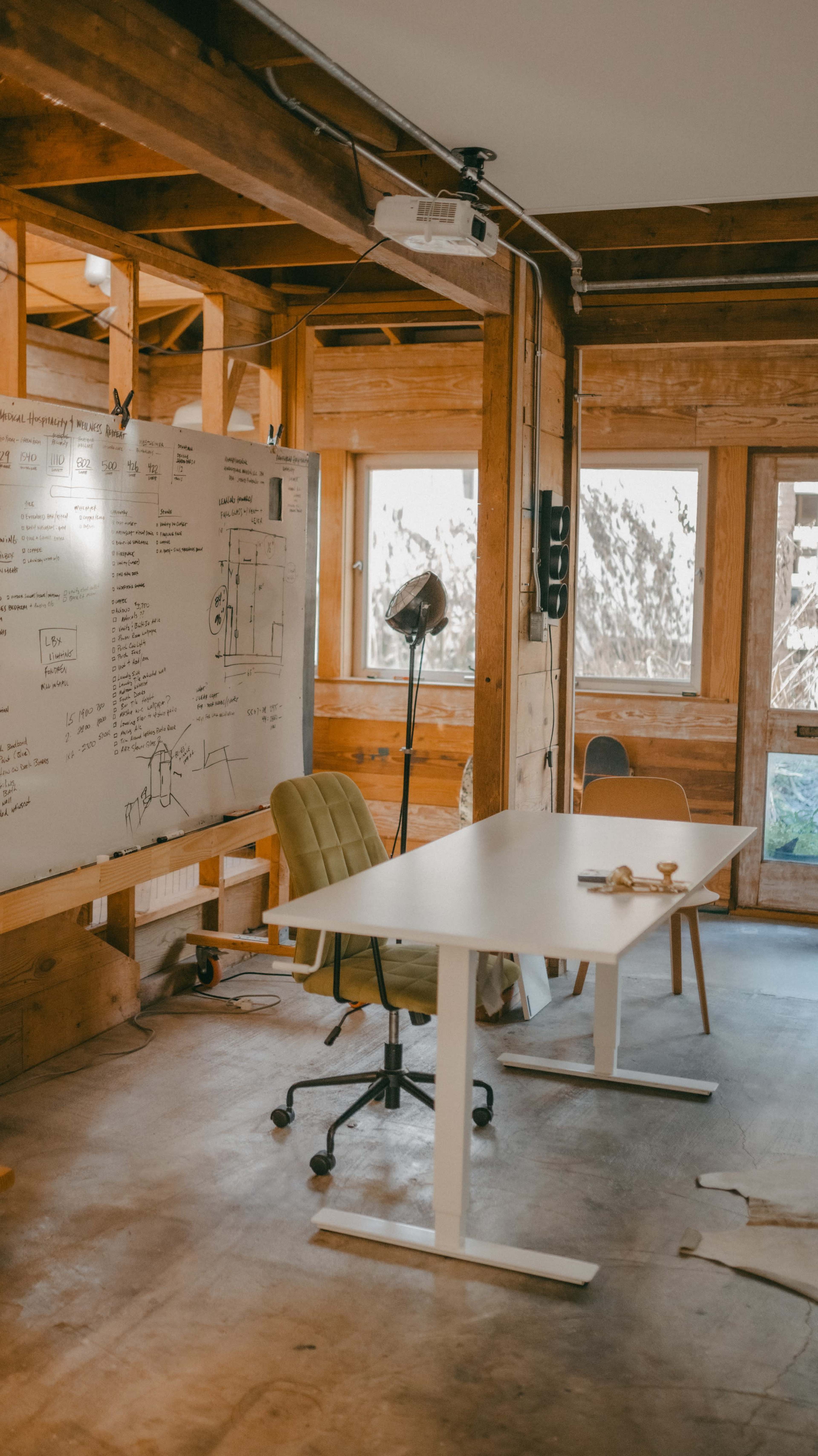 The image shows a minimalist office space with a whiteboard covered in notes, a desk with a chair, and natural light coming through large windows.