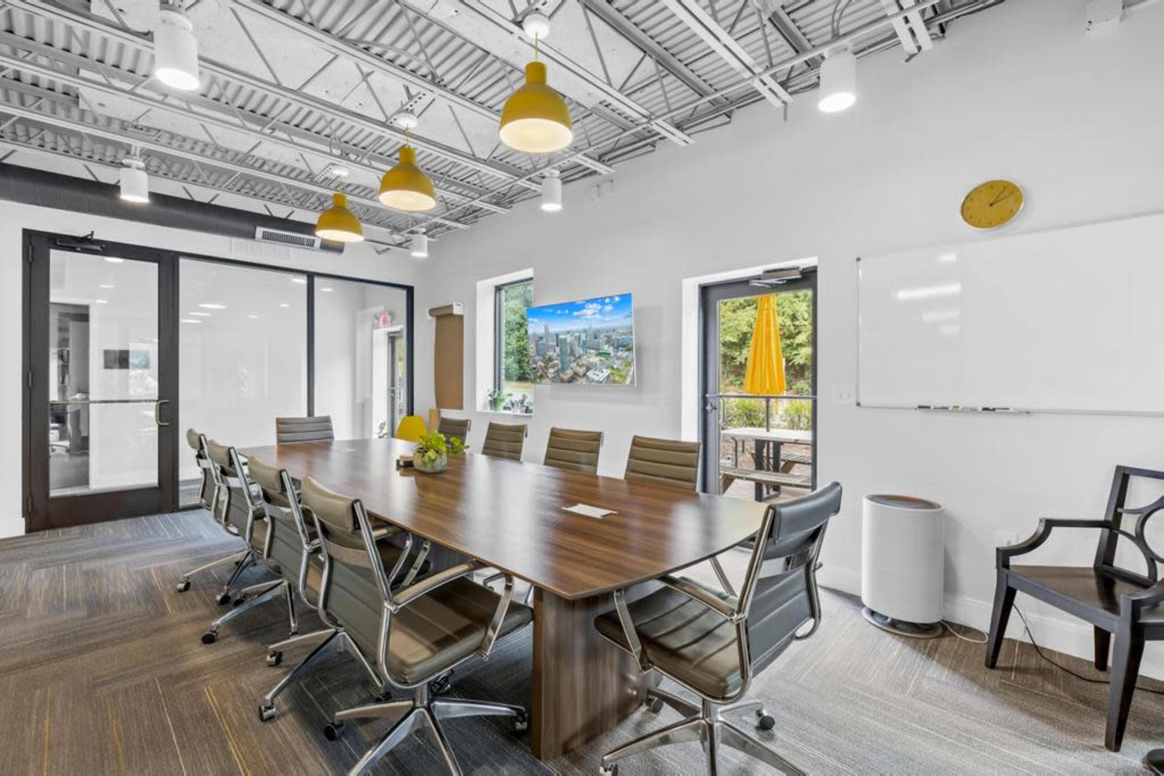A conference room features a large wooden table surrounded by gray office chairs, with windows showcasing a scenic view and a television mounted on the wall.