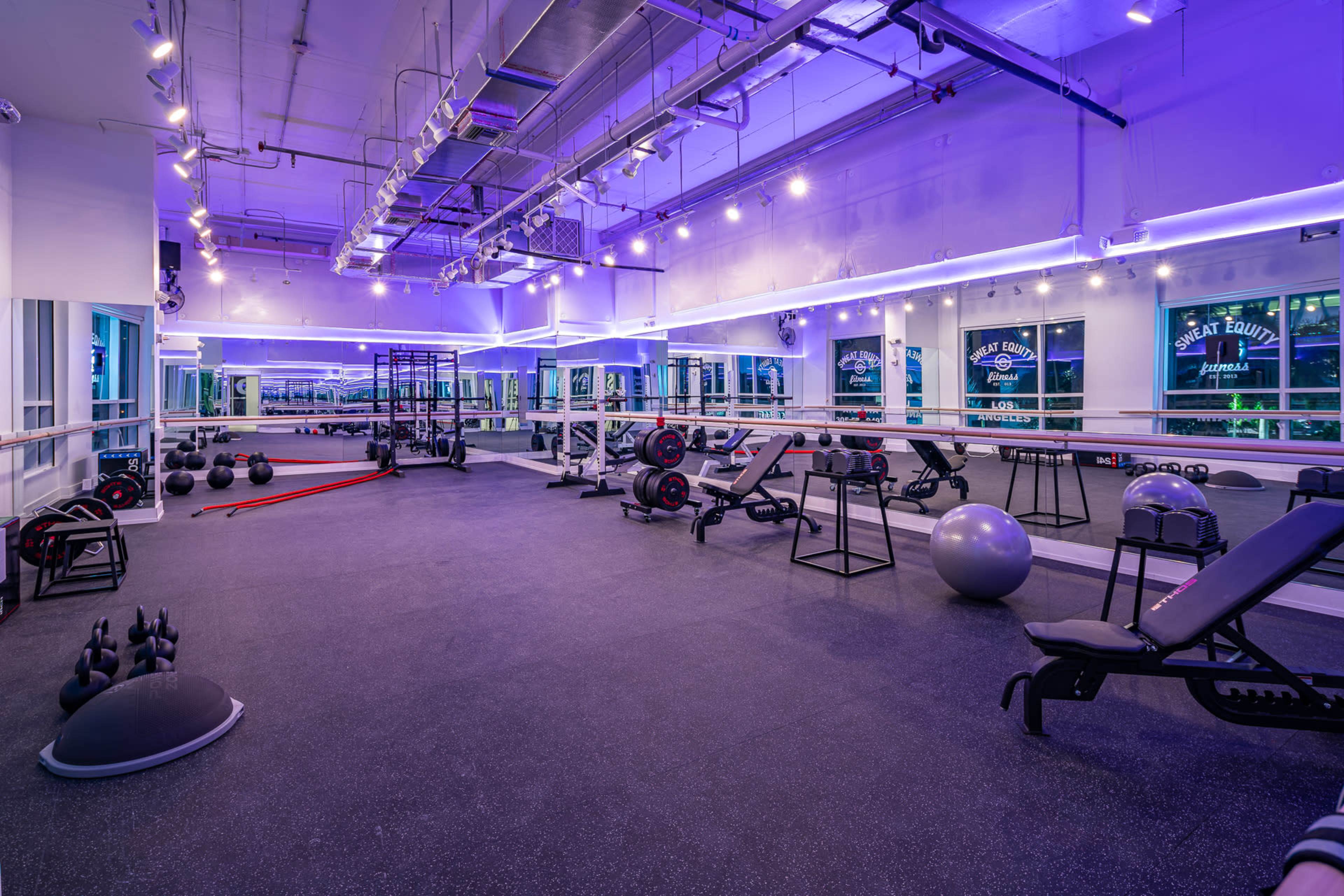 A well-equipped fitness studio featuring exercise machines, free weights, and various fitness accessories, illuminated by purple lights.