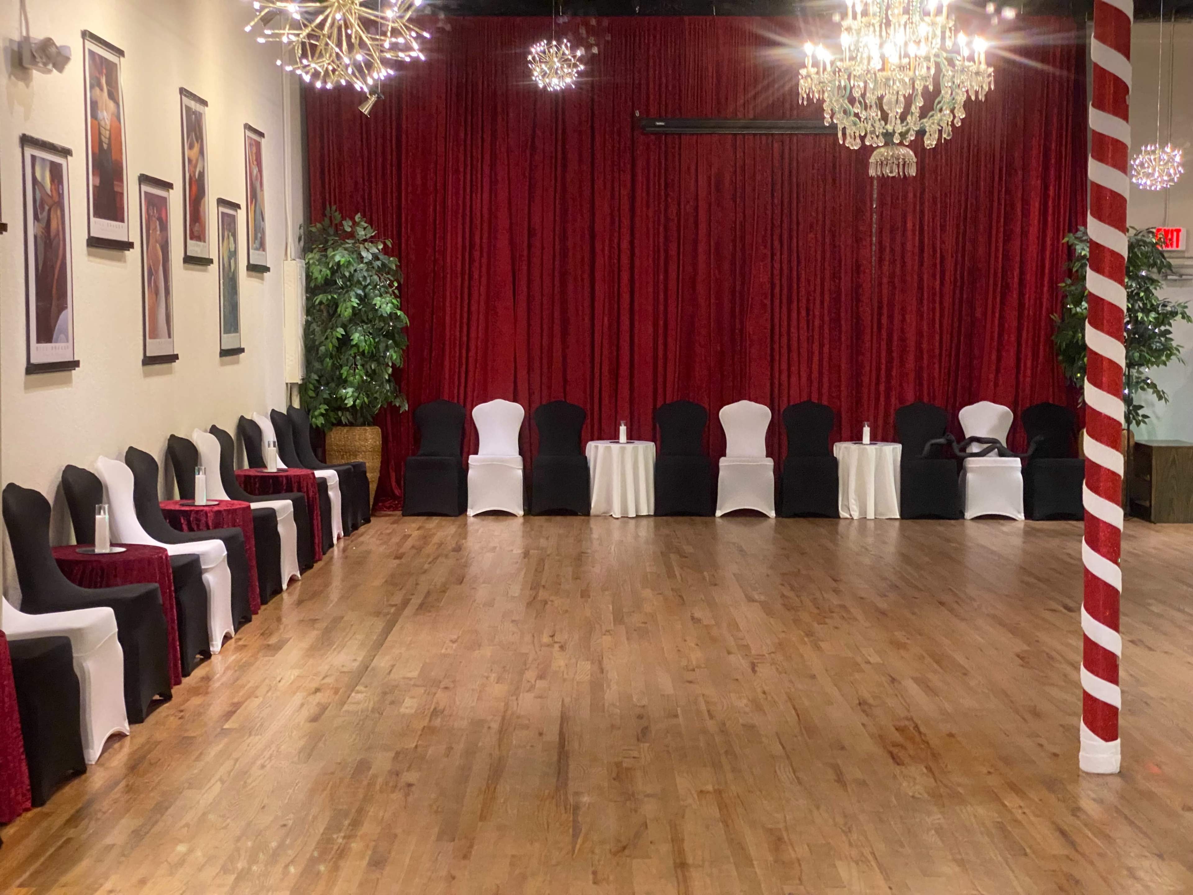 A dance hall with a wooden floor, decorated with black and white chairs arranged along the walls, and red curtains hanging behind them.
