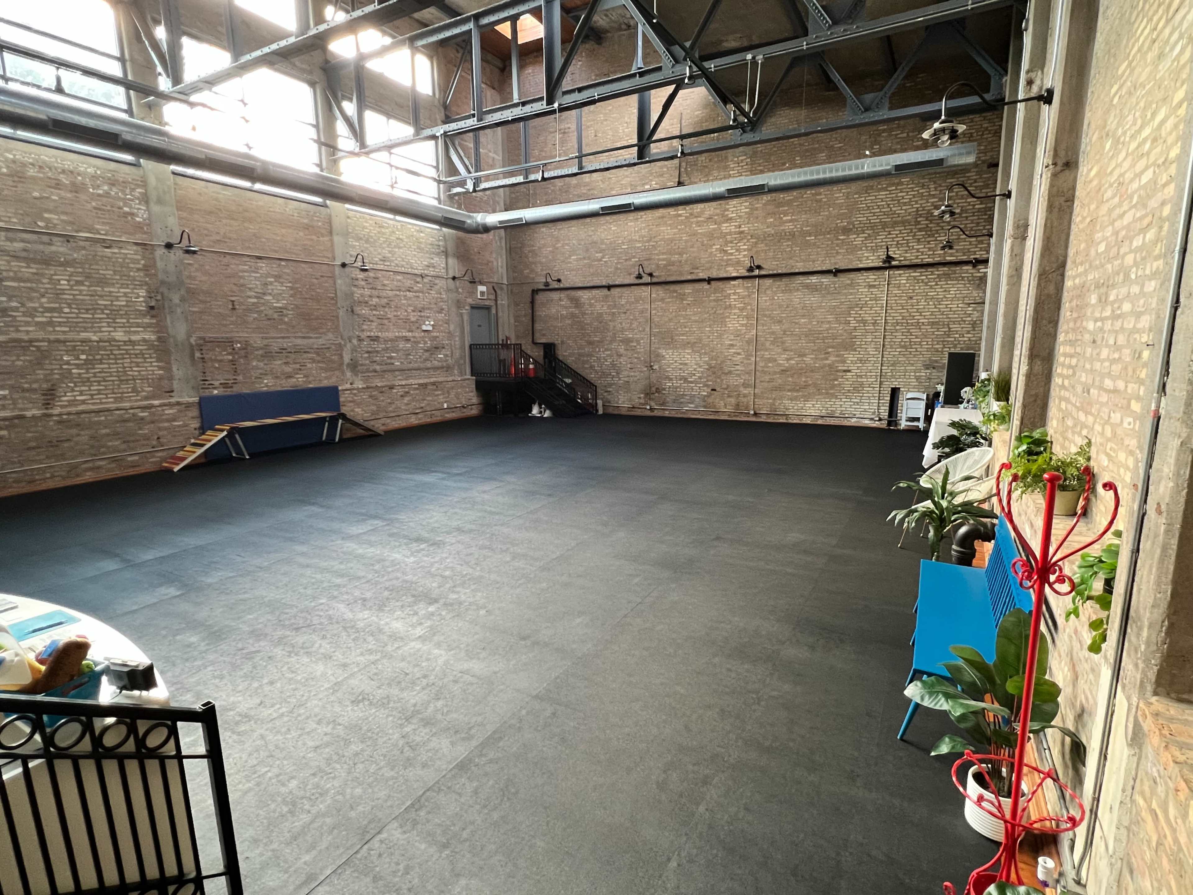 A spacious, empty indoor area with black rubber flooring and exposed brick walls, featuring a few pieces of furniture and plants along the edges.